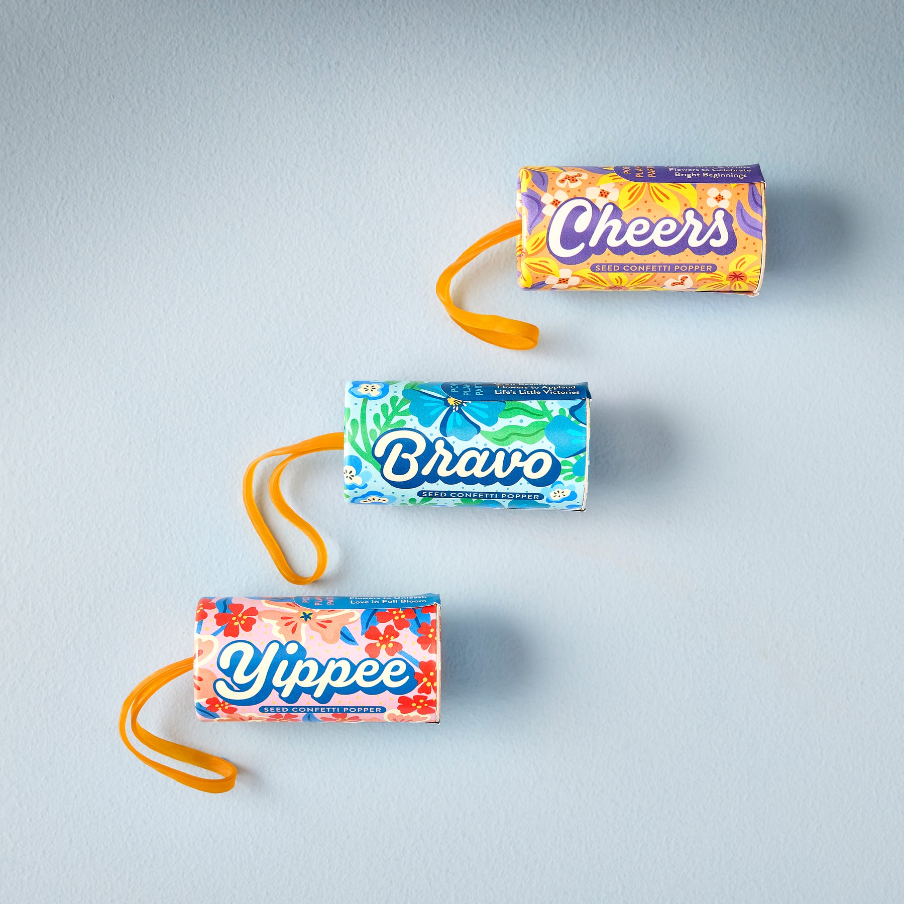 Colorful seed confetti poppers labeled "Cheers," "Bravo," and "Yippee" arranged on a light blue background.