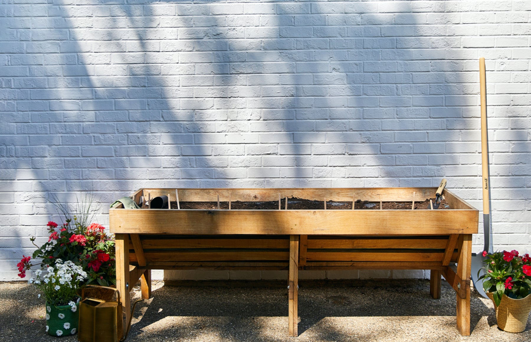 Wooden raised garden bed with soil, surrounded by colorful flowers and gardening tools against a white brick wall.