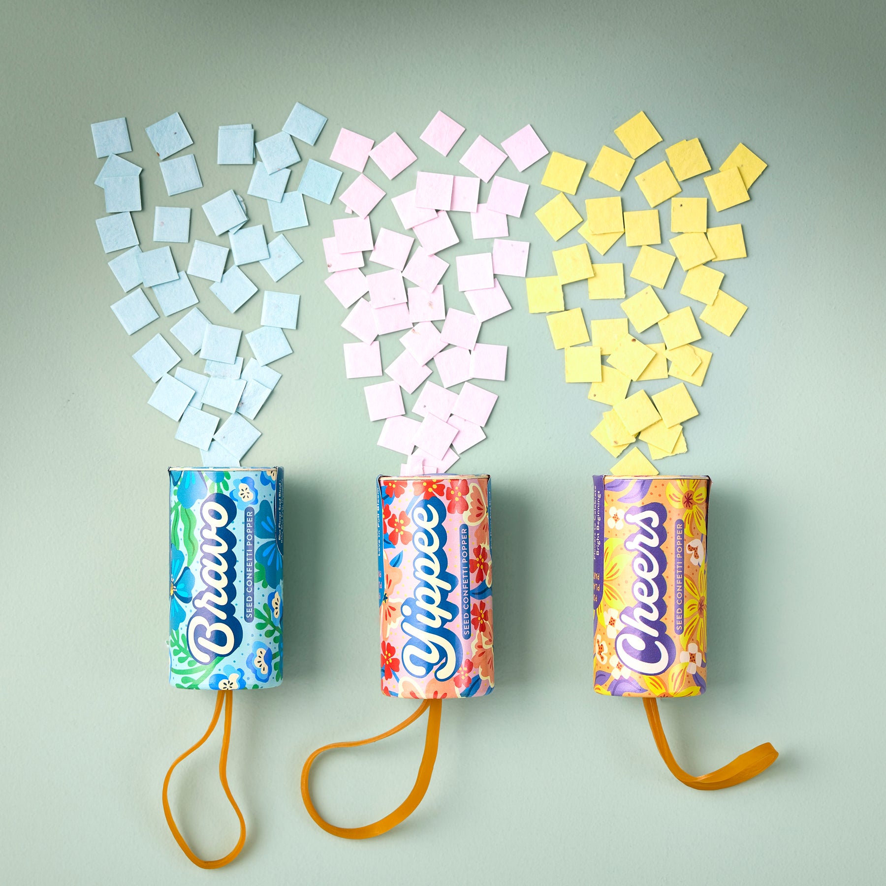 Colorful seed confetti poppers in blue, pink, and yellow, each releasing small square seed papers against a light background.