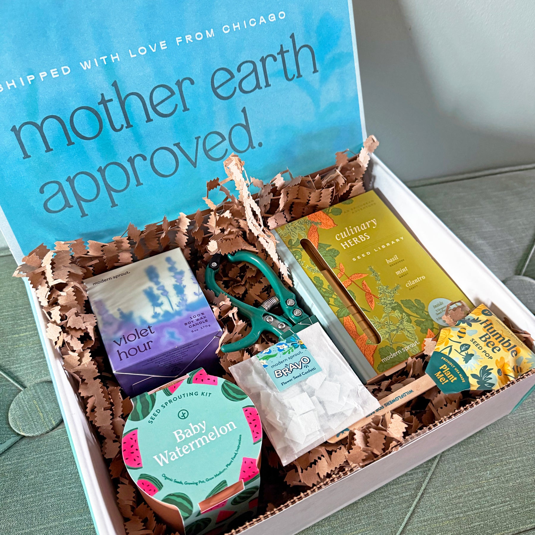 A gift box filled with gardening supplies, including seed packets, scissors, and a guidebook, on a textured surface.