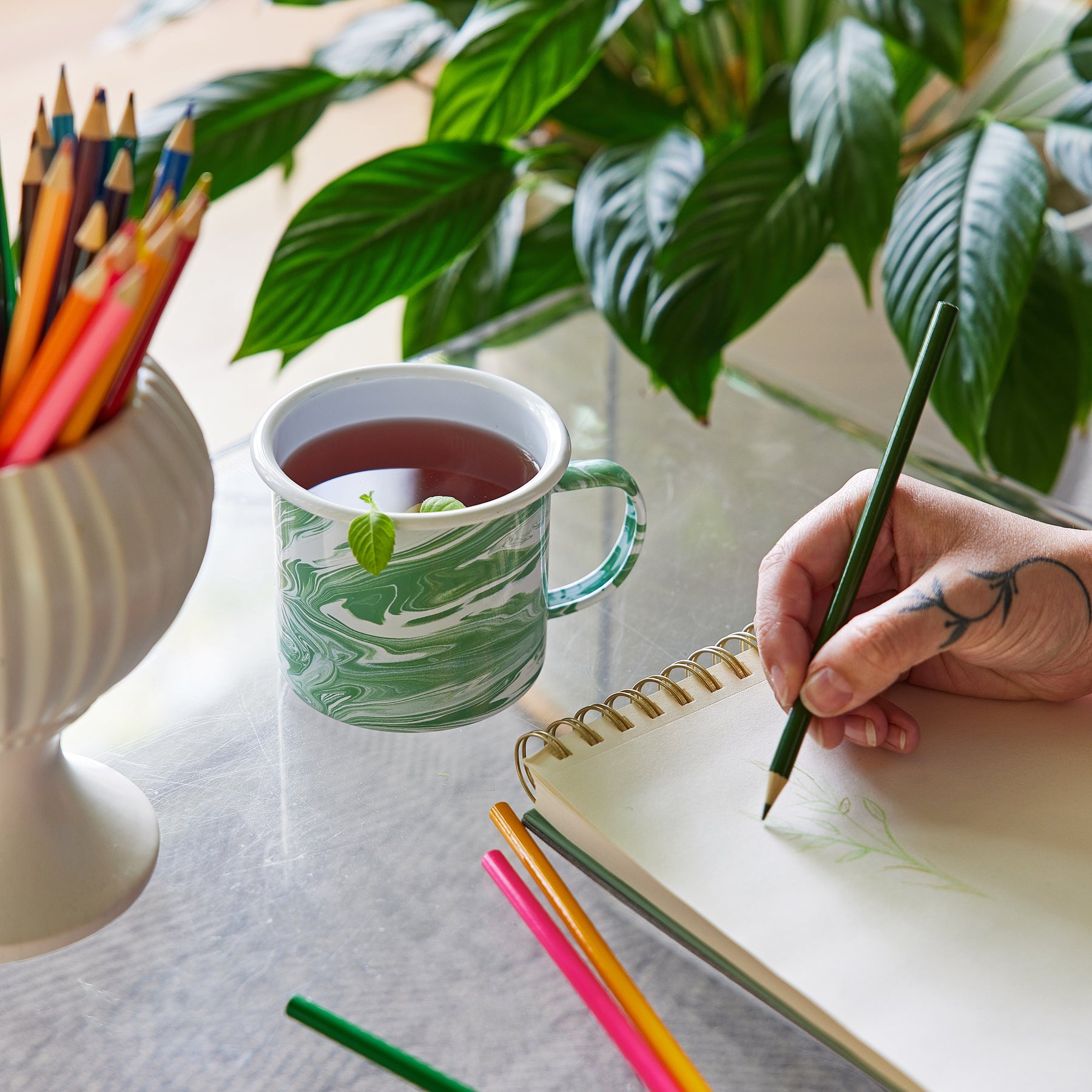 A green marbled mug filled with tea sits next to a sketchbook and colored pencils, with a hand drawing a leaf.