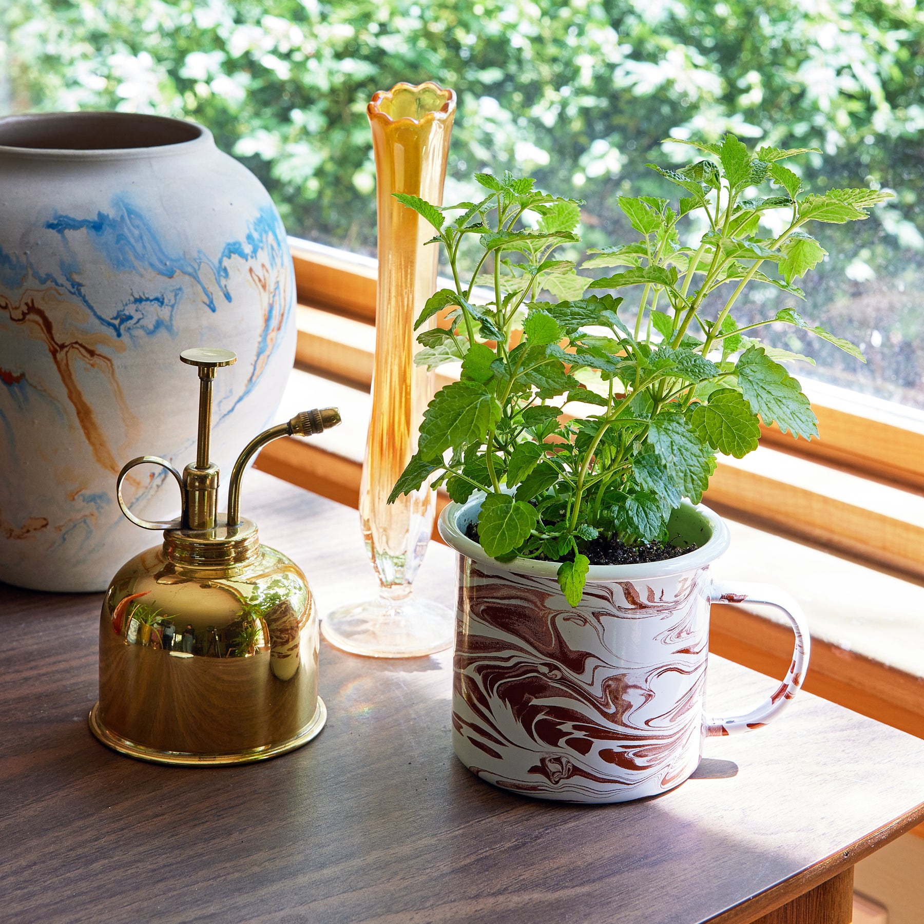 A marbled mug grow kit with lush green herbs sits on a wooden table beside a brass watering can and decorative vases.