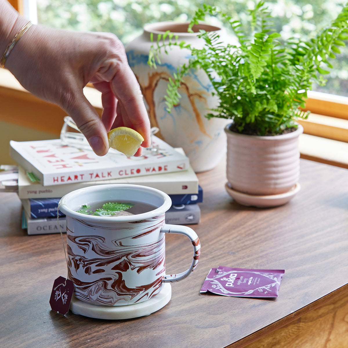 A hand adds a lemon slice to a marbled mug filled with herbal tea, surrounded by books and a potted fern.