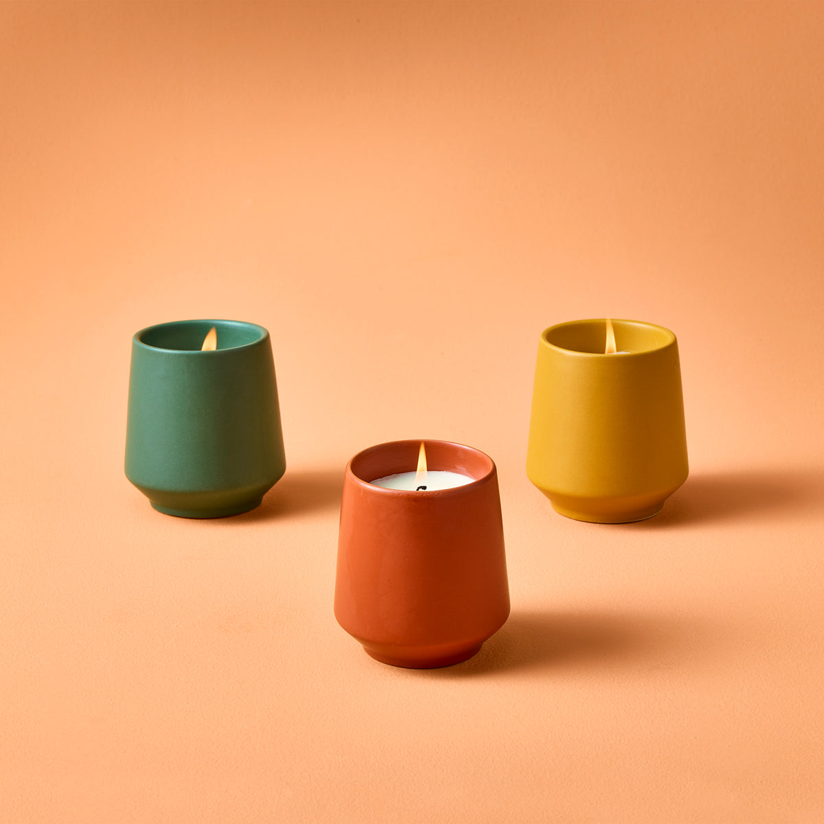 Three colorful candles in green, orange, and yellow ceramic holders, each with a lit wick, on a peach background.