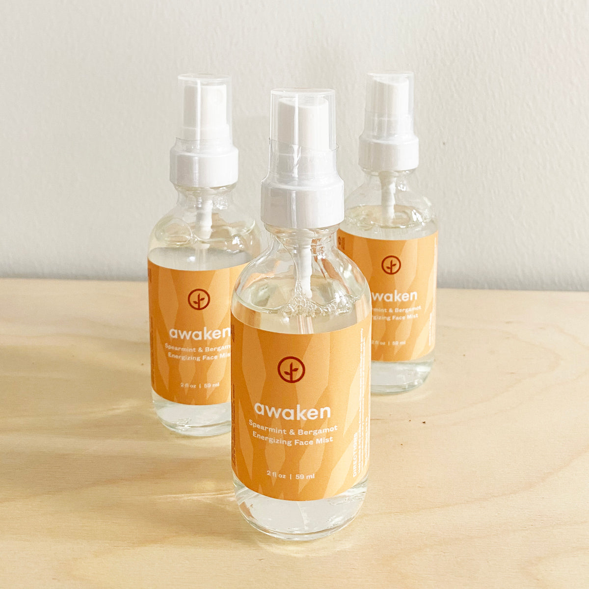 Three bottles of Shine Bright Face Mist, featuring orange labels with the word "awaken," arranged on a wooden surface.