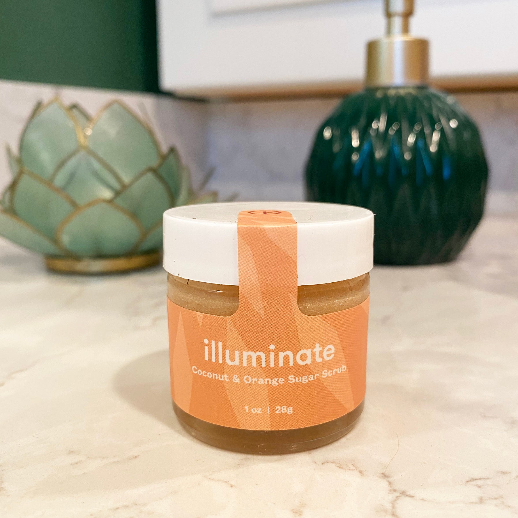Three jars of Shine Bright Sugar Scrub in coconut and orange scents, displayed on a marble countertop with decorative elements.