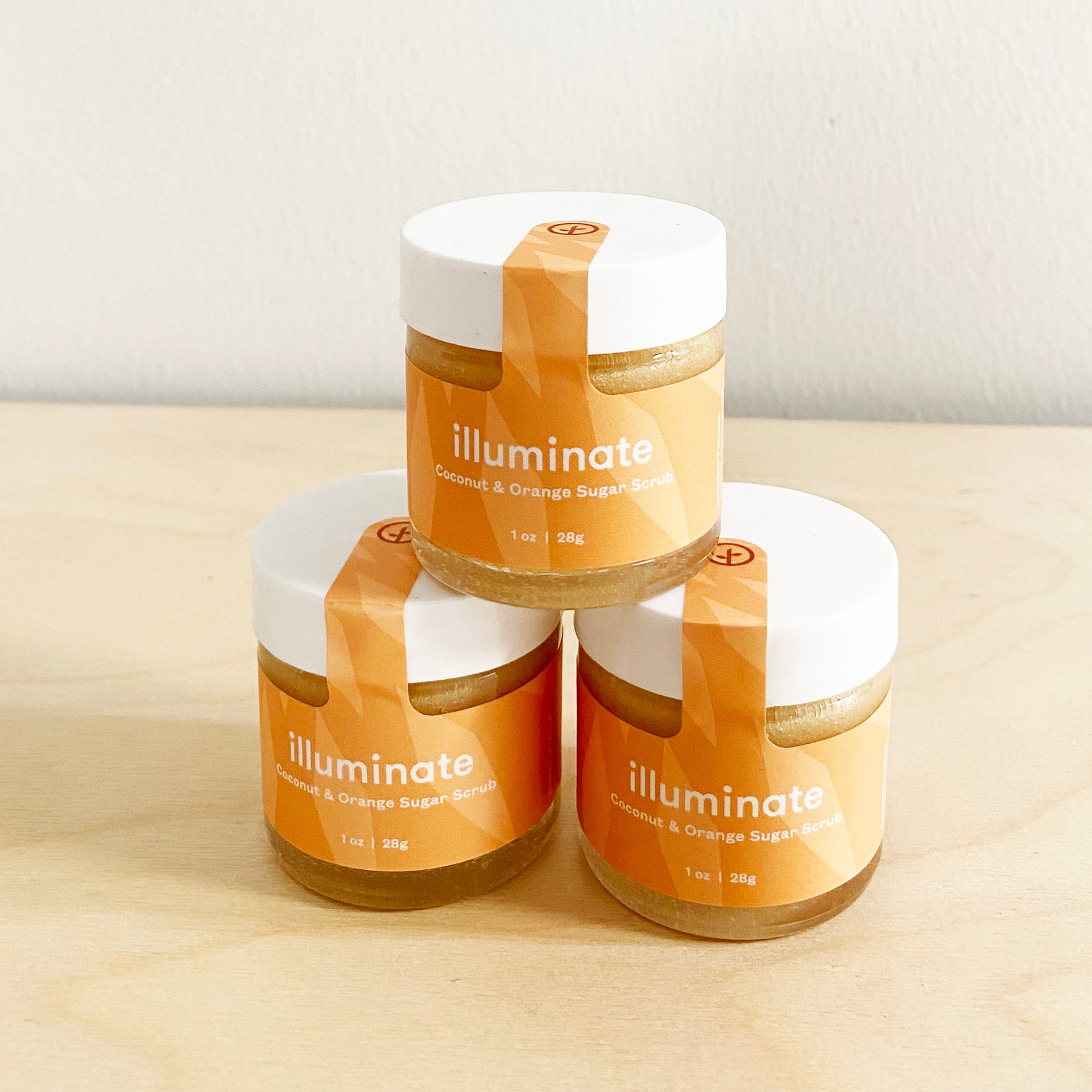 Three jars of Shine Bright Sugar Scrub in coconut and orange scent, stacked on a wooden surface.