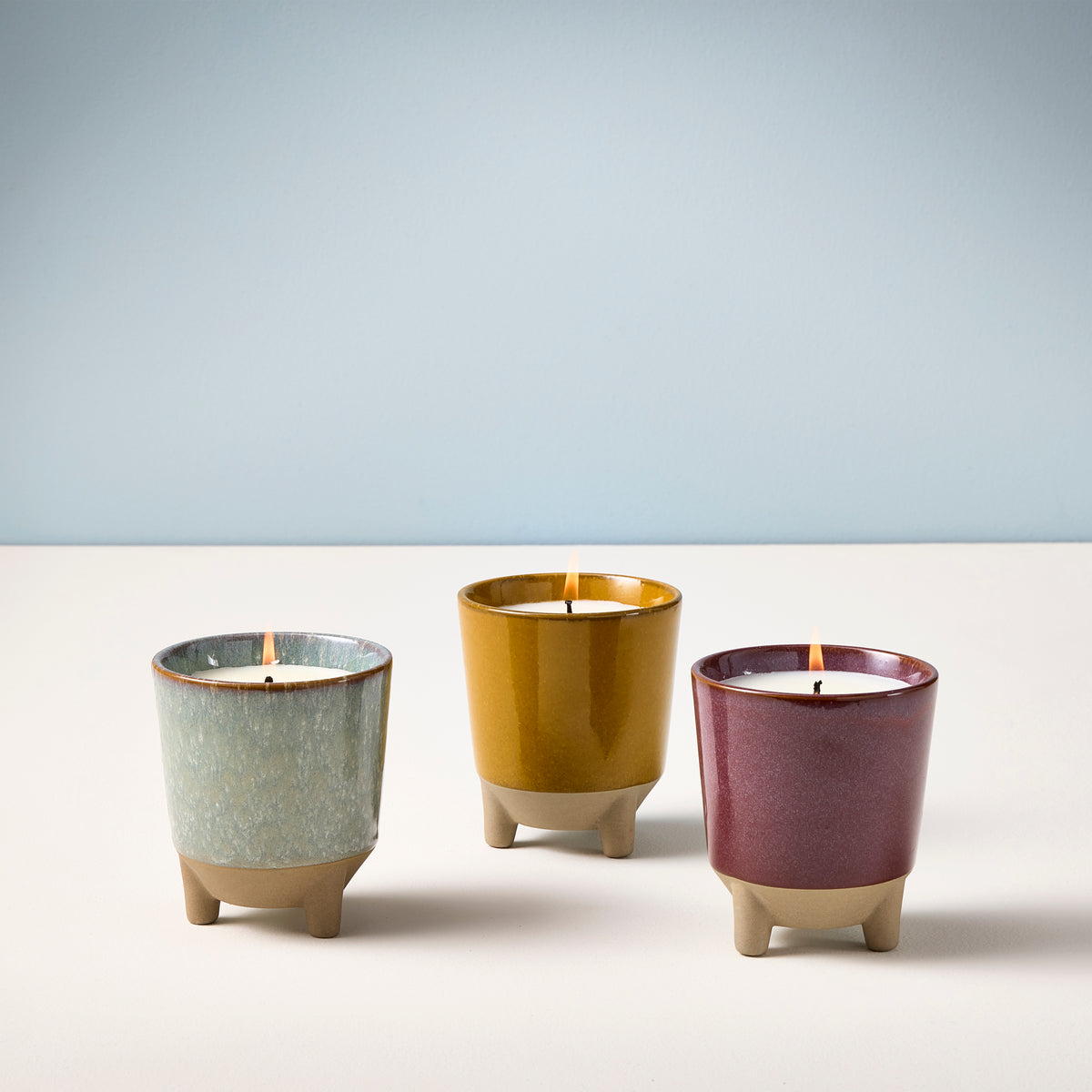 Three colorful candles in ceramic pots with legs, glowing softly against a light blue background.