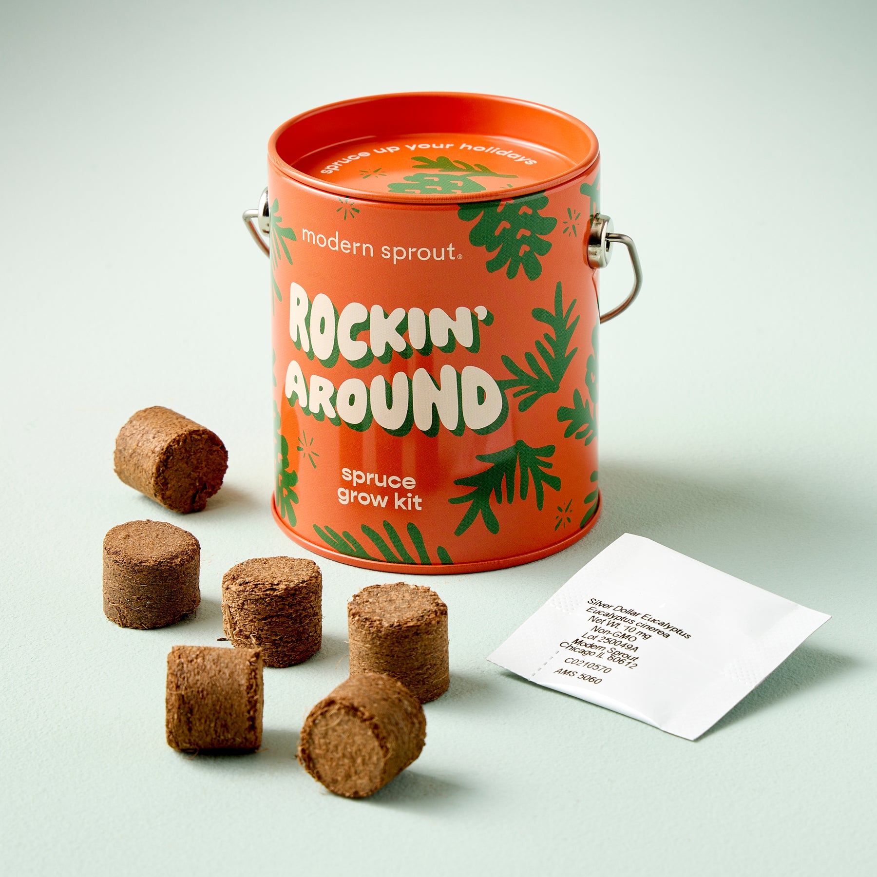 Bright orange tin labeled "Rockin' Around" containing a spruce grow kit, with soil pellets arranged around it.