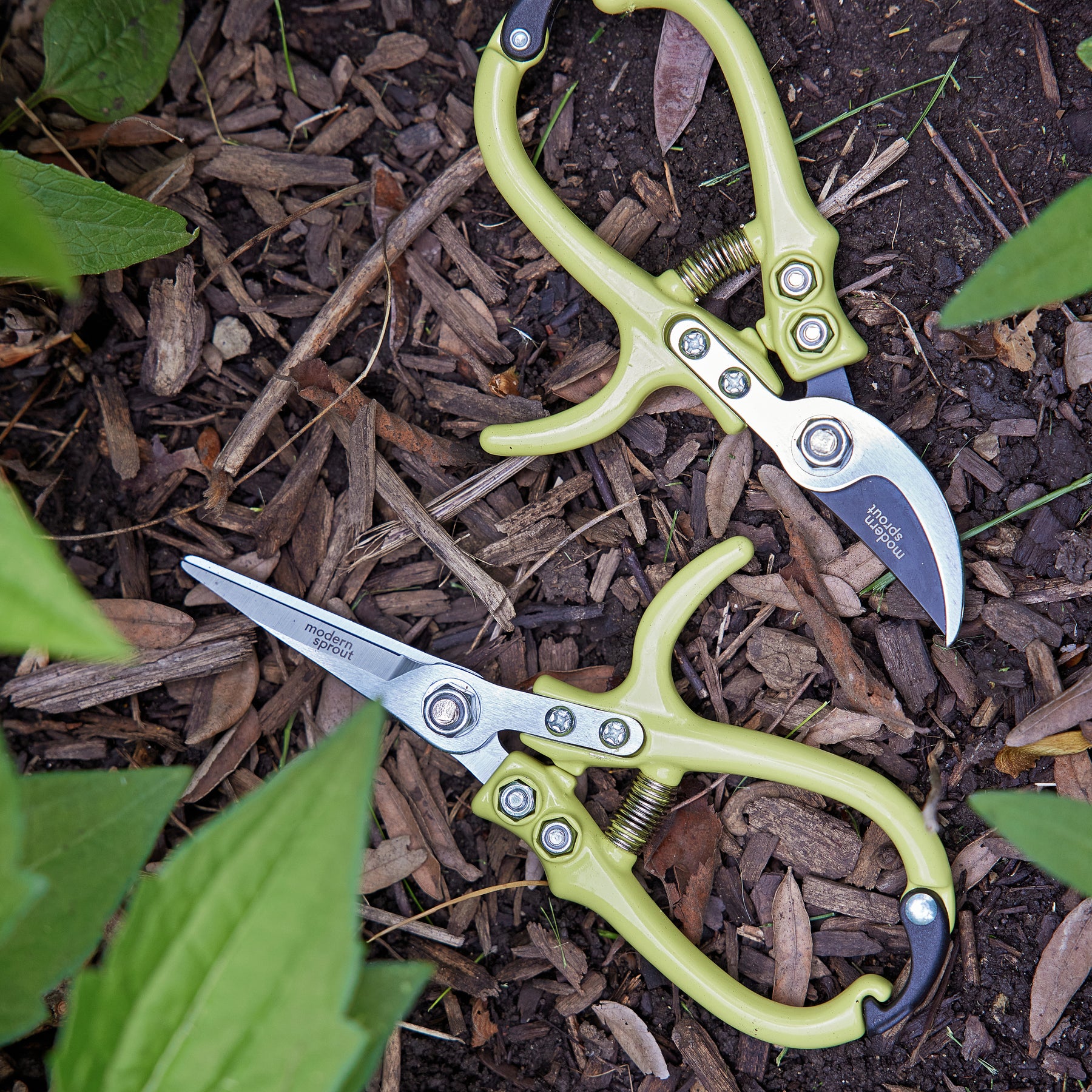 Gardening shears and pruners with green handles resting on mulch among plants, showcasing sharp stainless steel blades.