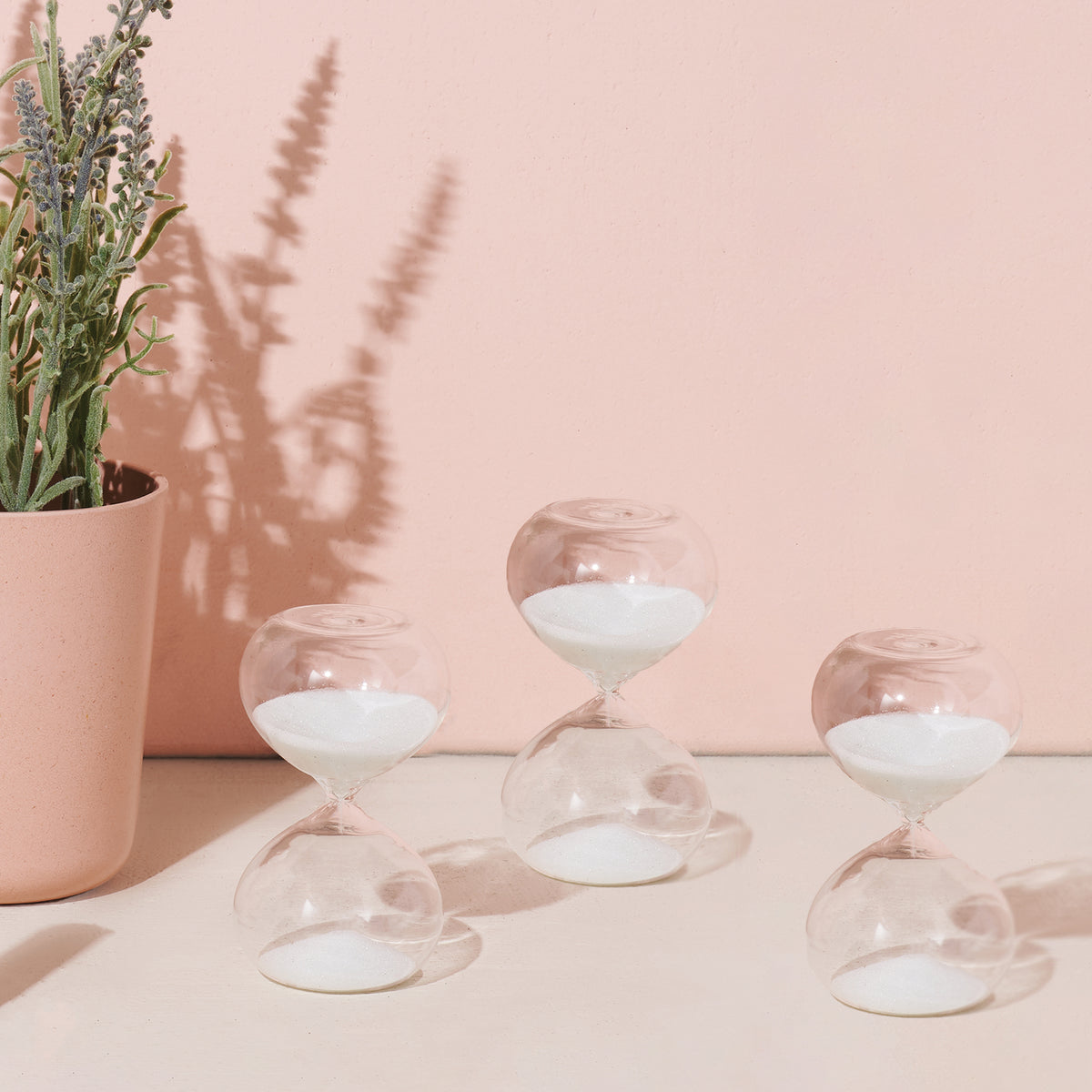 Three glass hourglasses with white sand, placed on a light surface beside a pink planter with lavender.
