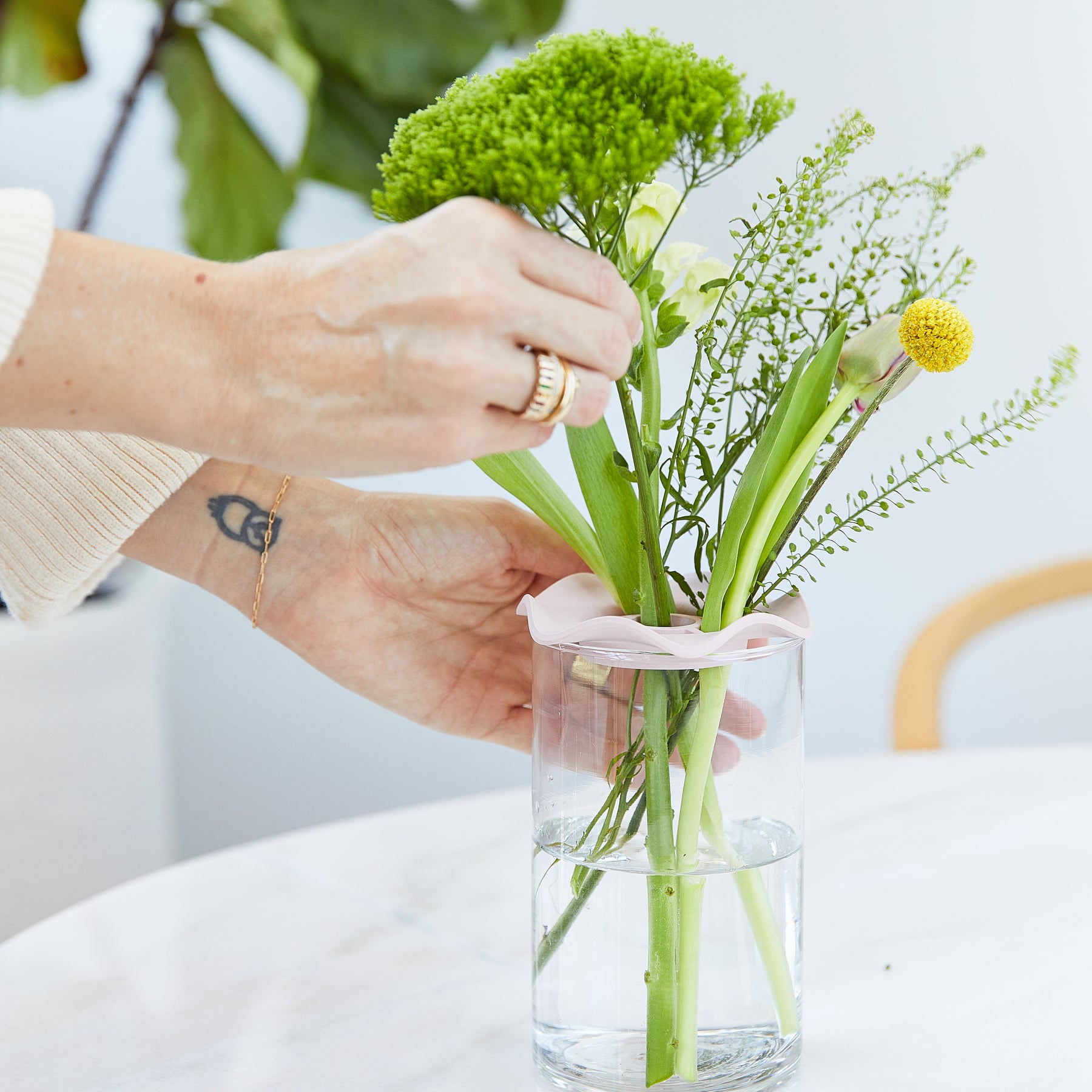 A hand adjusts flowers in a glass vase using a pink Botanical Keeper, showcasing floral arrangement support.