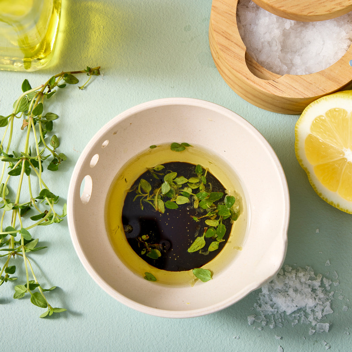 A ceramic dish filled with olive oil and fresh herbs, surrounded by lemon slices, sea salt, and a wooden container.