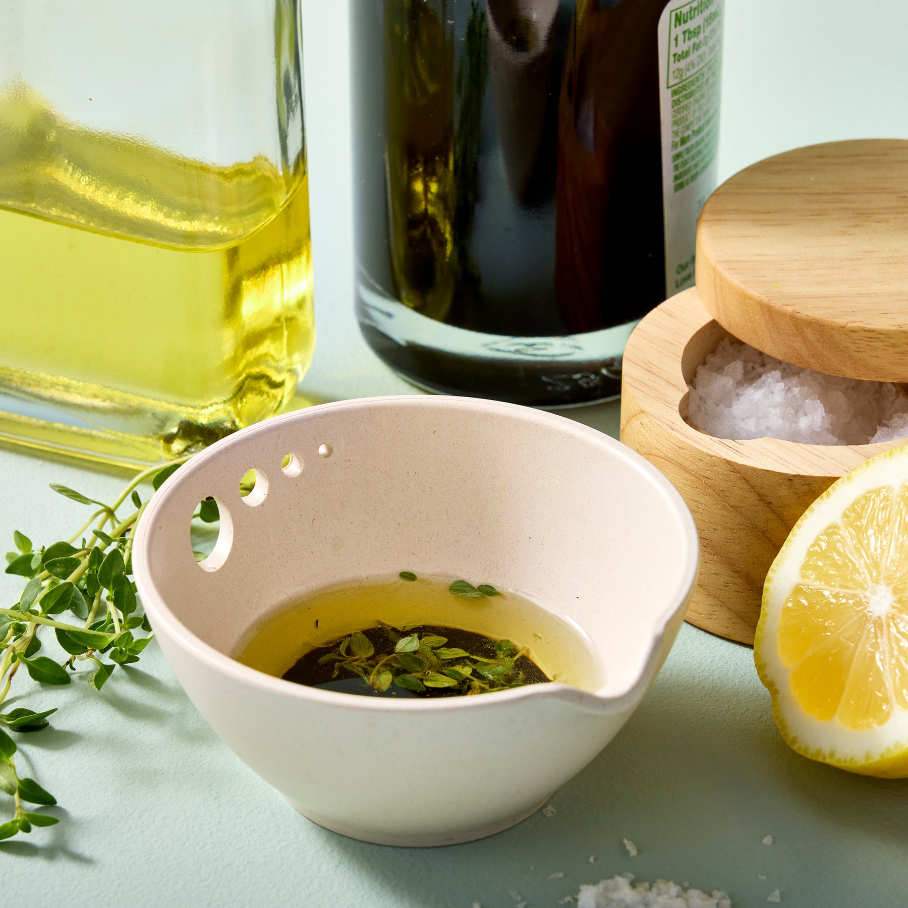 A beige prep dish with herbs and oil, surrounded by olive oil, salt, and lemon, showcasing a modern kitchen setup.