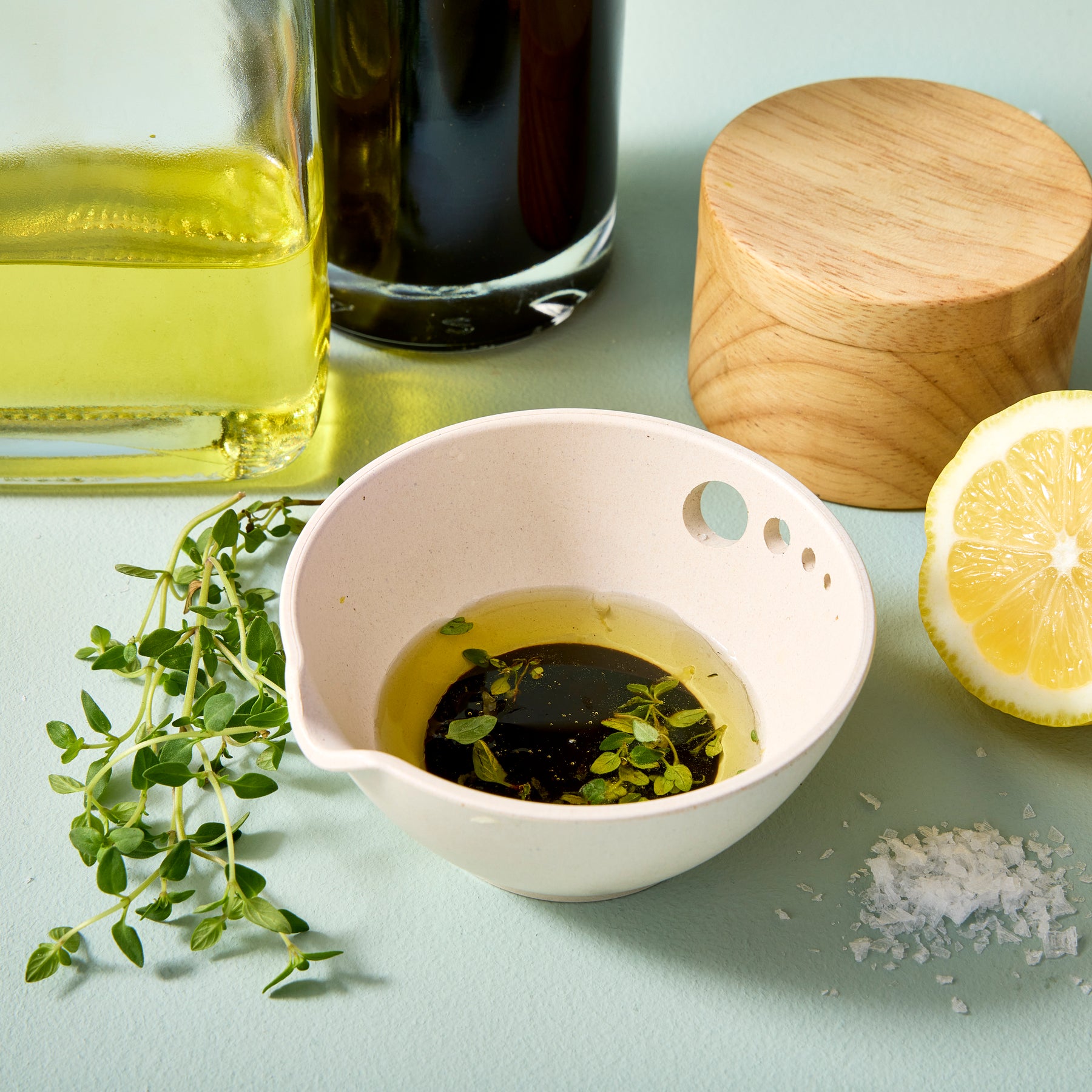 A light-colored prep dish with olive oil, fresh thyme, and lemon slices, surrounded by salt and a wooden lid.