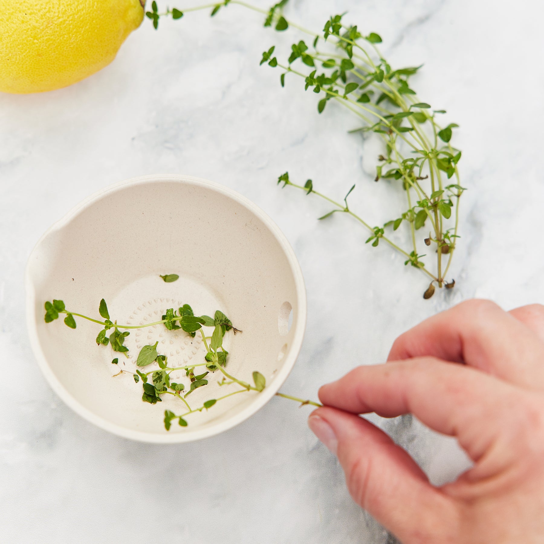 A hand holds a sprig of thyme above a beige dish designed for peeling, grating, and prepping ingredients, with a lemon nearby.