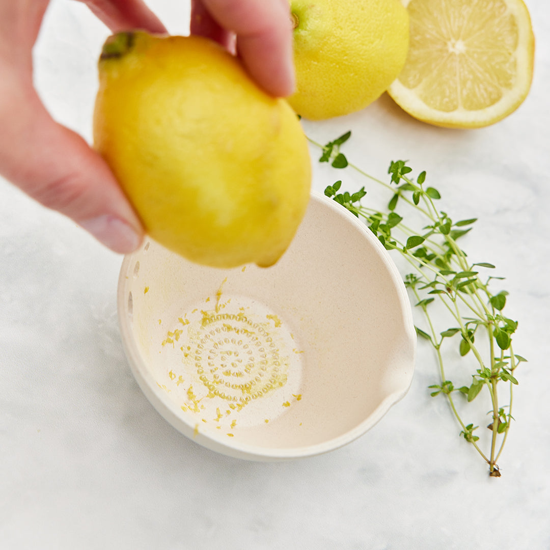 A hand holds a lemon above a ceramic dish with zest inside, surrounded by fresh lemons and thyme sprigs.
