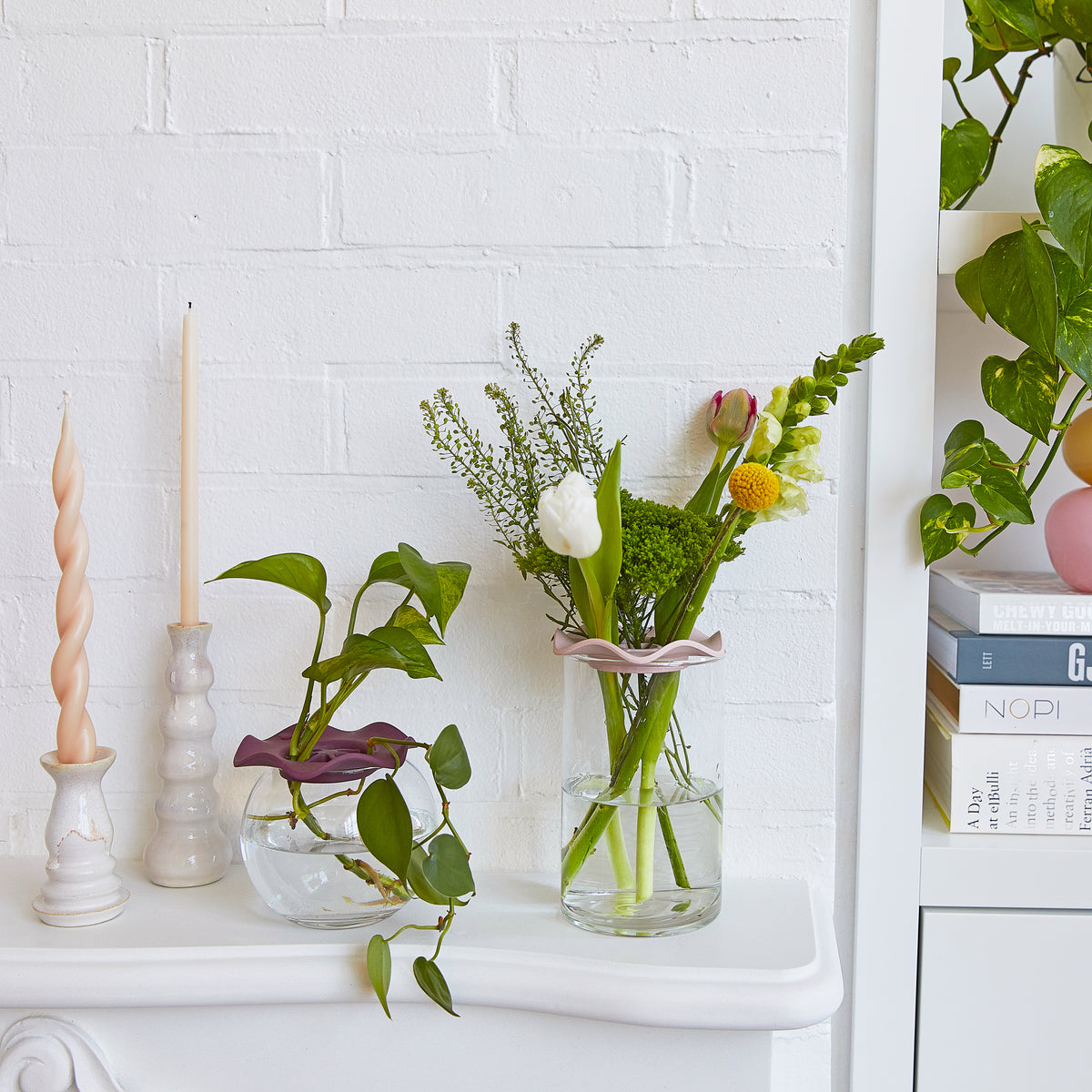 Two Botanical Keepers holding fresh flowers in vases on a white shelf, alongside decorative candles and plants.