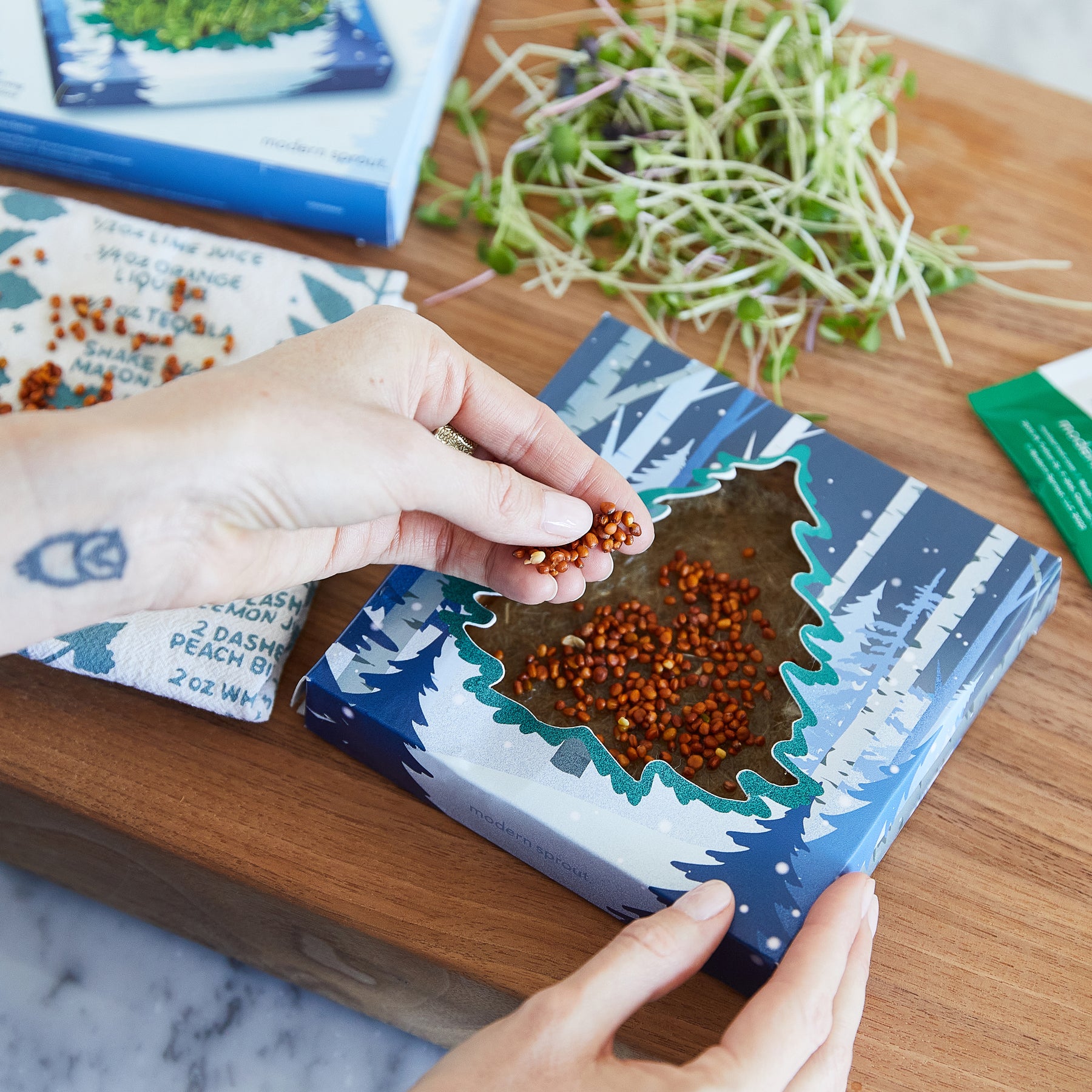 A hand sprinkles seeds from a box featuring a tree design, surrounded by fresh microgreens on a wooden surface.