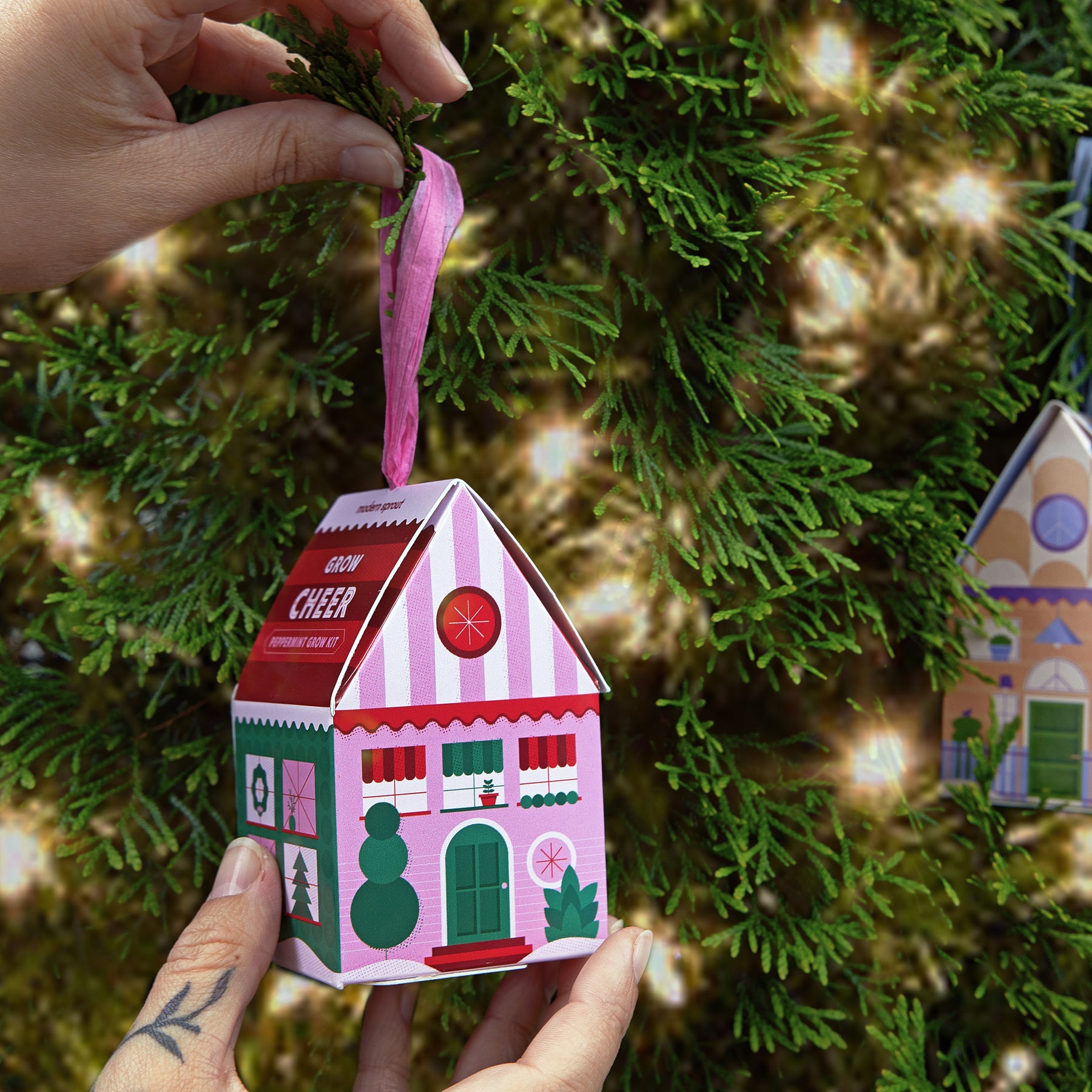 Colorful holiday ornament shaped like a gingerbread house, being hung on a Christmas tree branch.