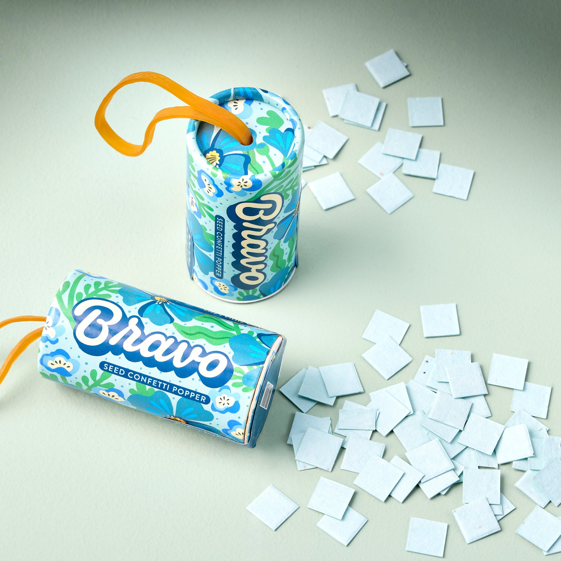 Colorful seed confetti poppers with blue floral design, surrounded by scattered seed paper squares on a light green background.