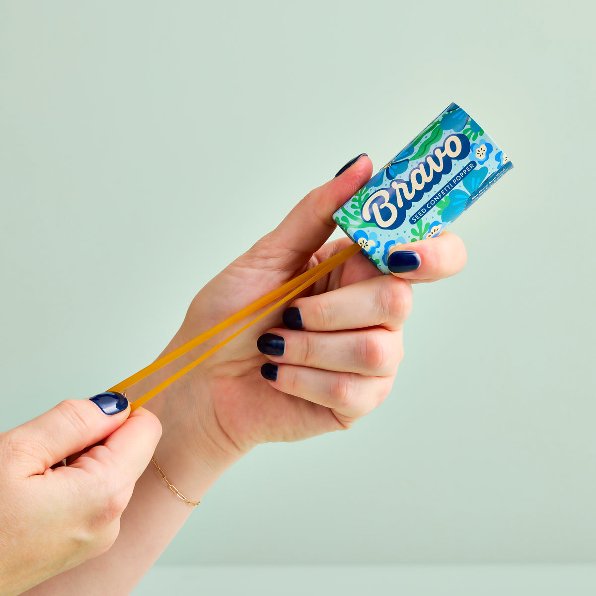 A hand holds a colorful seed confetti popper, pulling back the elastic band ready to launch seeds.