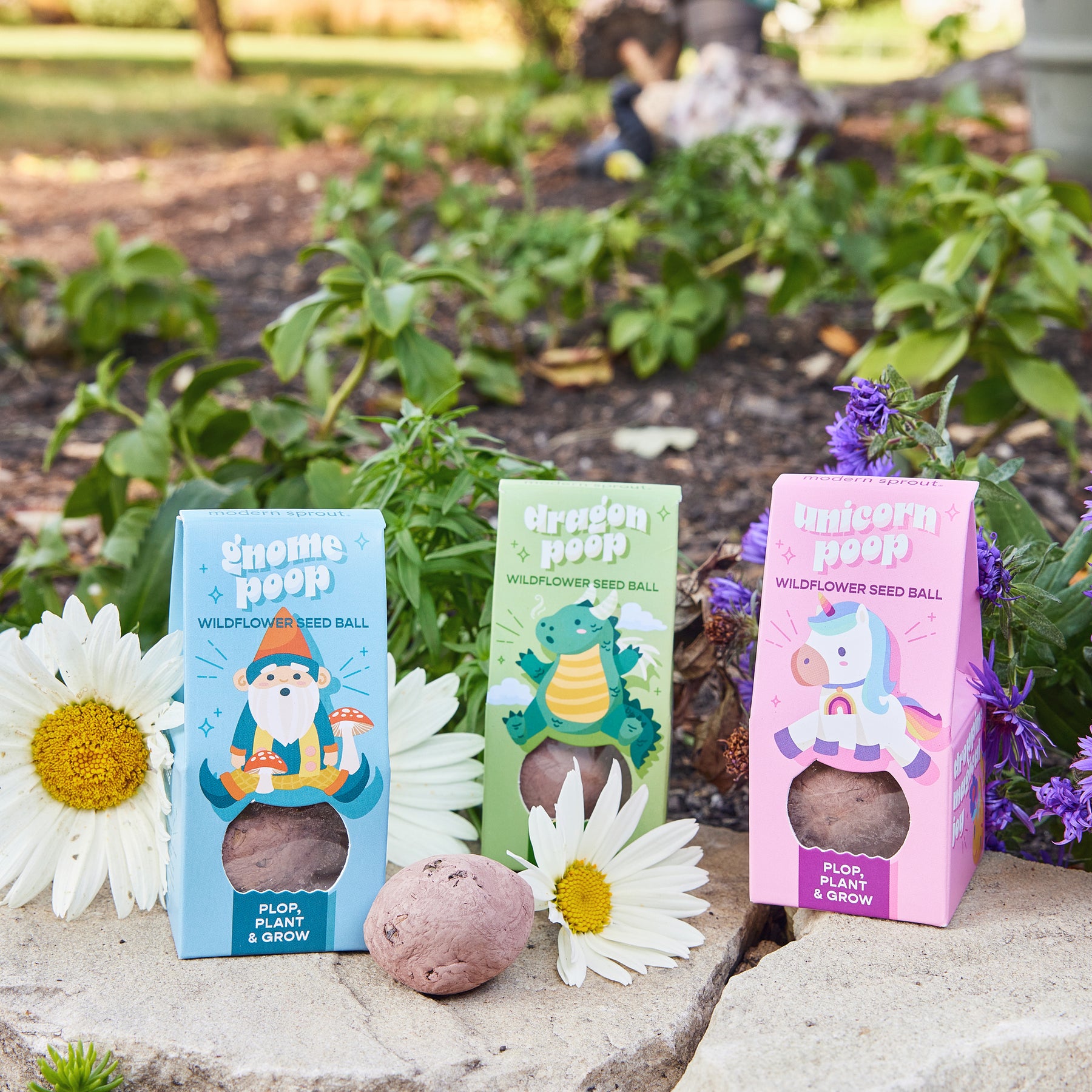 Three colorful seed ball boxes labeled Gnome Poop, Dragon Poop, and Unicorn Poop, surrounded by flowers and greenery.