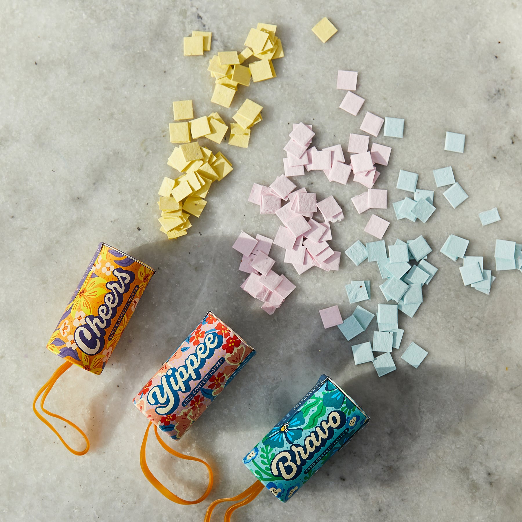 Colorful seed confetti poppers in vibrant packaging, surrounded by pastel seed confetti pieces on a marble surface.
