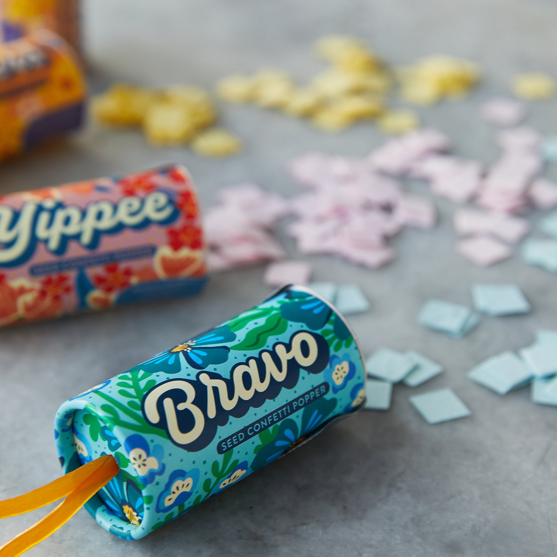 Colorful seed confetti poppers in vibrant packaging, surrounded by scattered pastel confetti pieces on a gray surface.