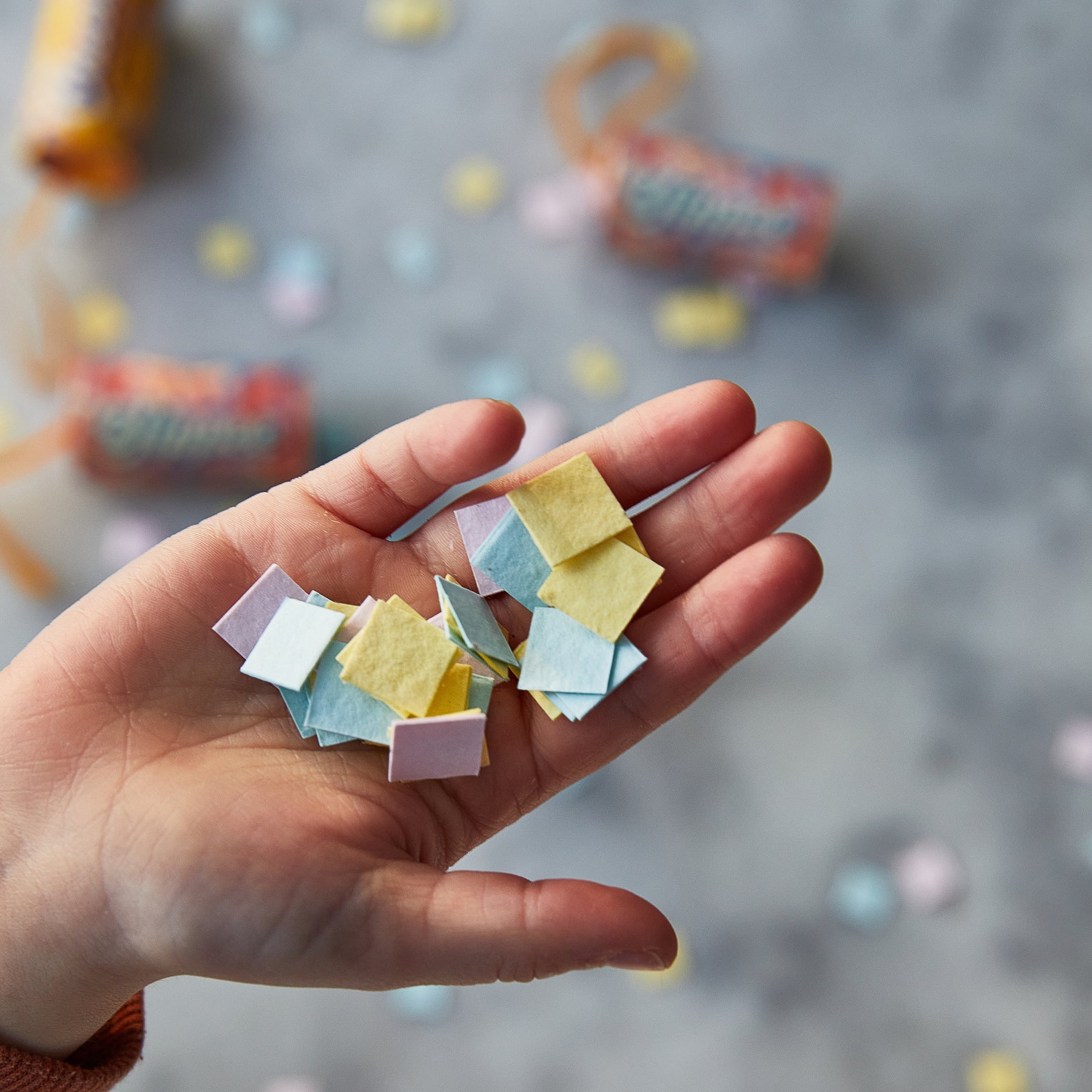 A hand holds colorful seed confetti pieces, with additional confetti scattered in the background.