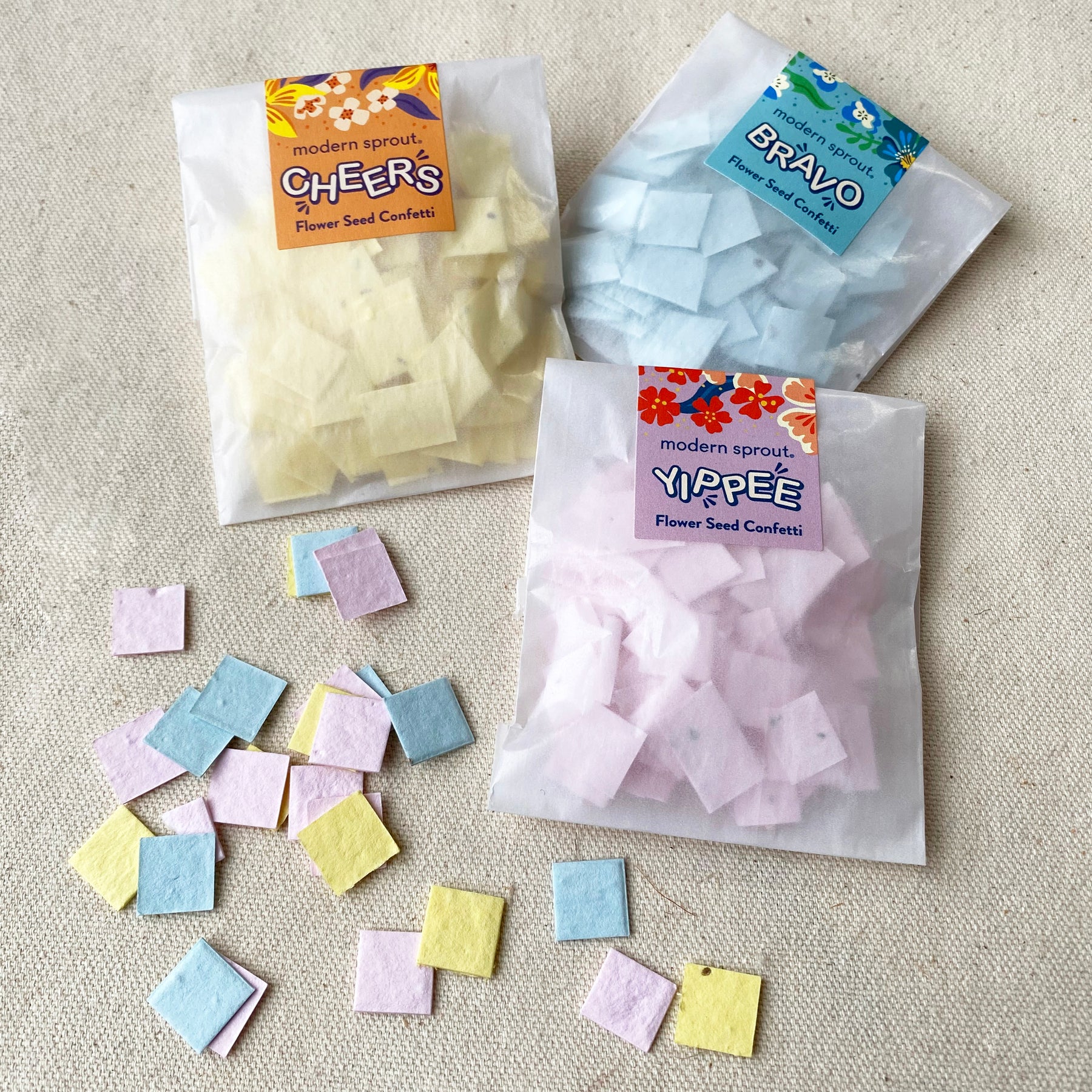 Colorful seed paper confetti refill packs in yellow, blue, and pink, scattered on a light fabric surface.