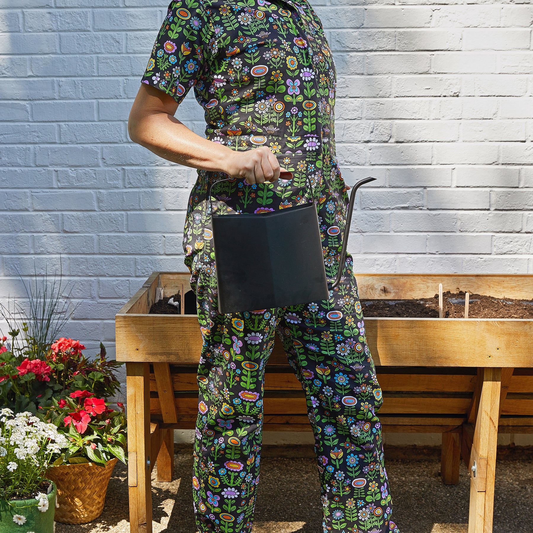 A person in a colorful floral outfit holds a sleek black watering can next to a wooden planter filled with soil.