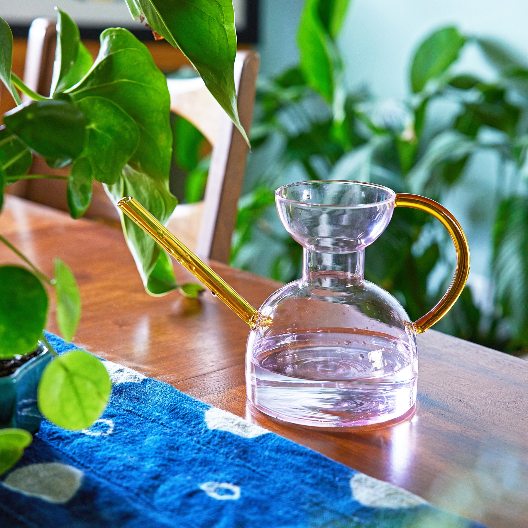 Clear glass watering can with a yellow handle, resting on a wooden table surrounded by green plants.