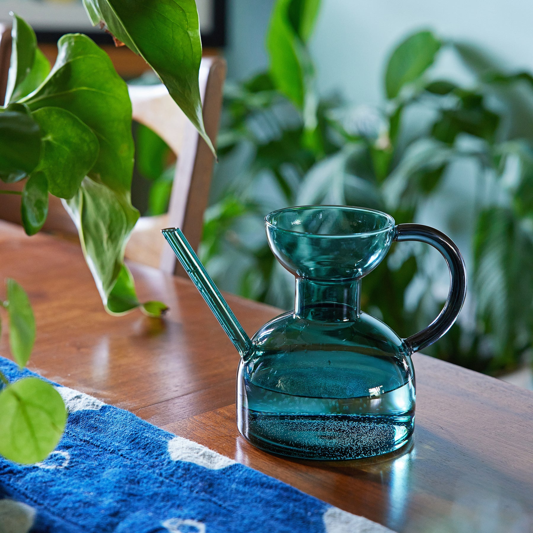 Elegant glass watering can with a curved spout, placed on a wooden table surrounded by greenery.