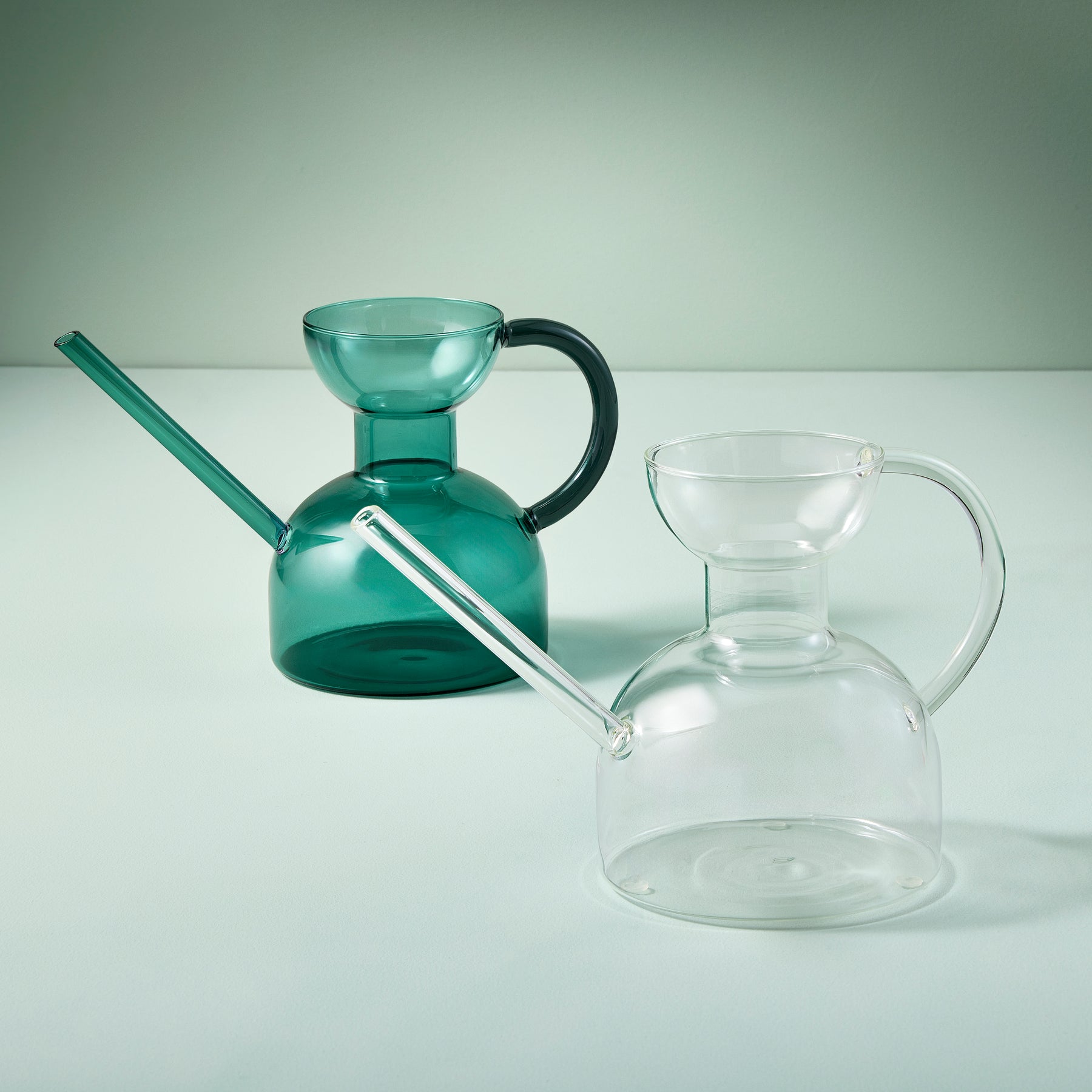 Two elegant glass watering cans in green and clear, featuring unique shapes and long spouts for precise watering.