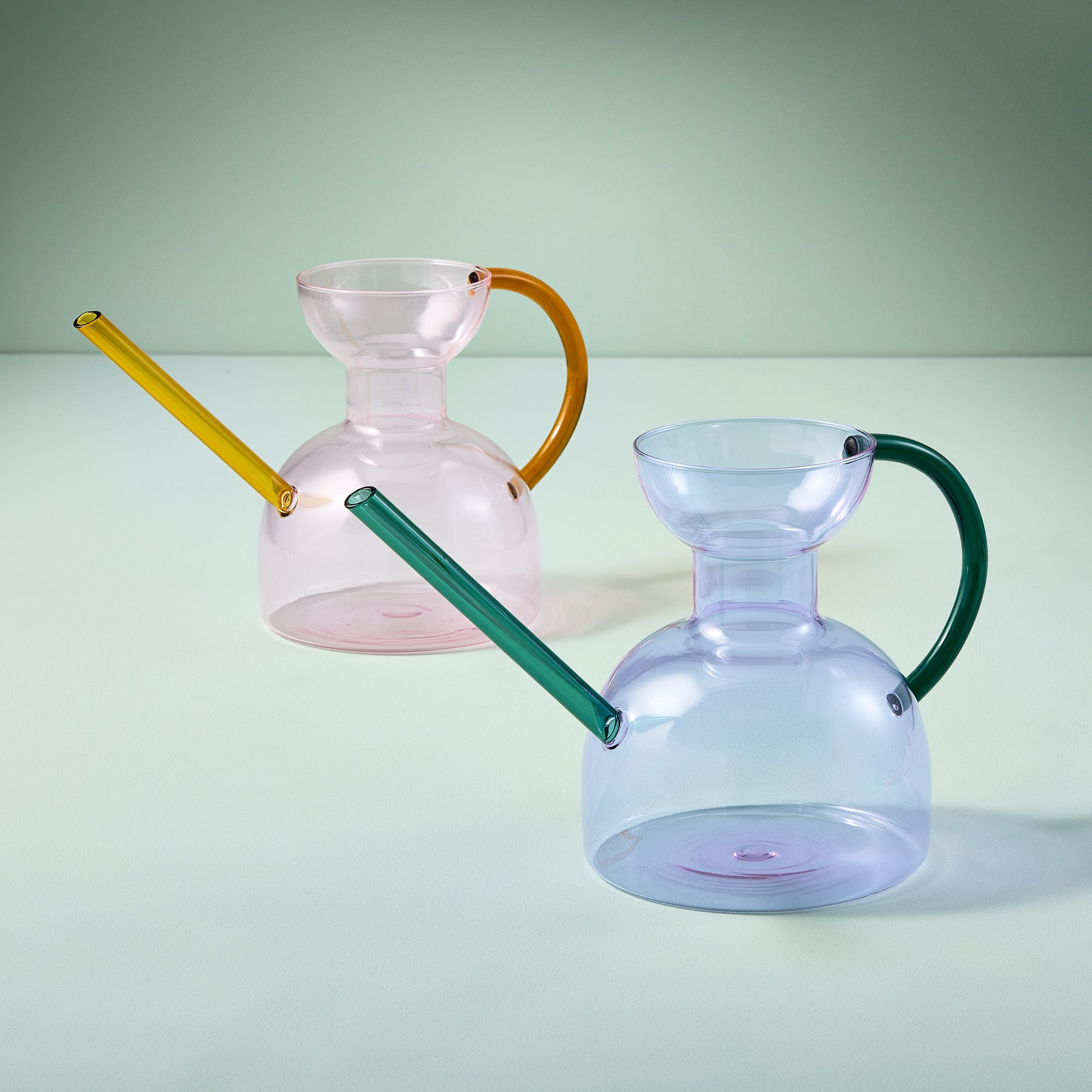 Two stylish glass watering cans in pink and blue, featuring colorful handles, set against a soft green background.