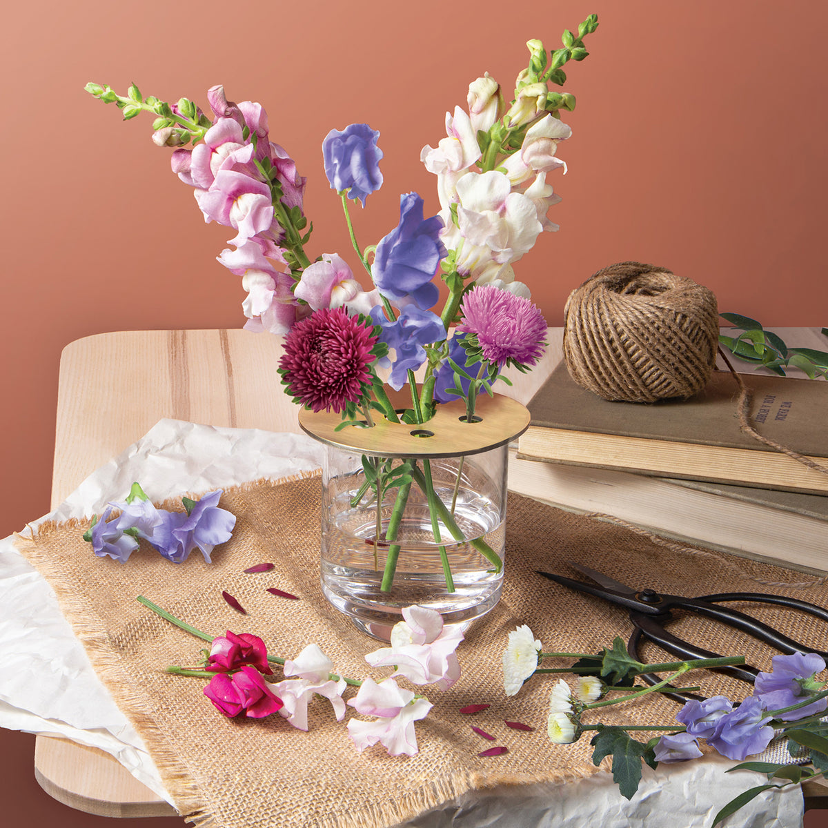 Brass flower frog holds vibrant flowers in a clear vase on a wooden table, surrounded by floral clippings and twine.