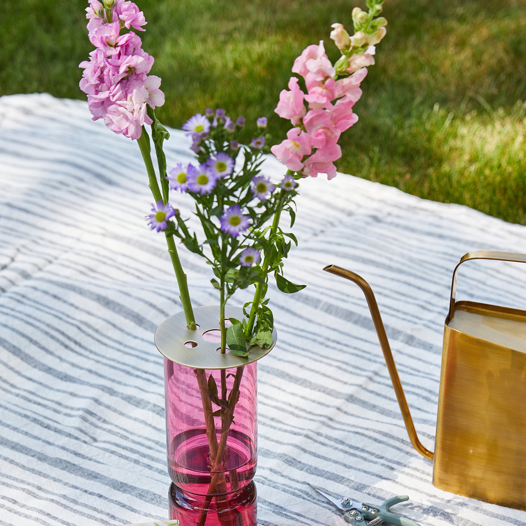 Brass flower frog atop a pink vase holding pink and purple flowers on a striped fabric outdoors.