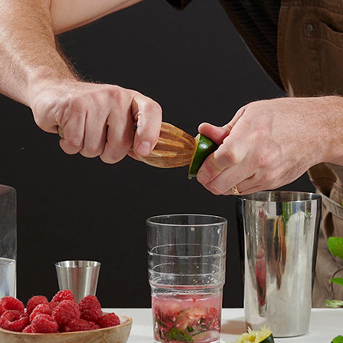 A person using a wooden citrus reamer to extract juice from a lime over a glass, with fresh raspberries and cocktail tools nearby.