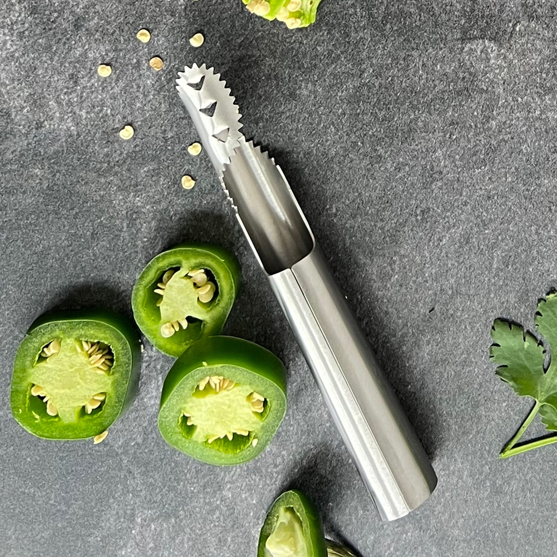 Stainless steel pepper de-seeder tool beside halved green peppers and scattered seeds on a dark surface.