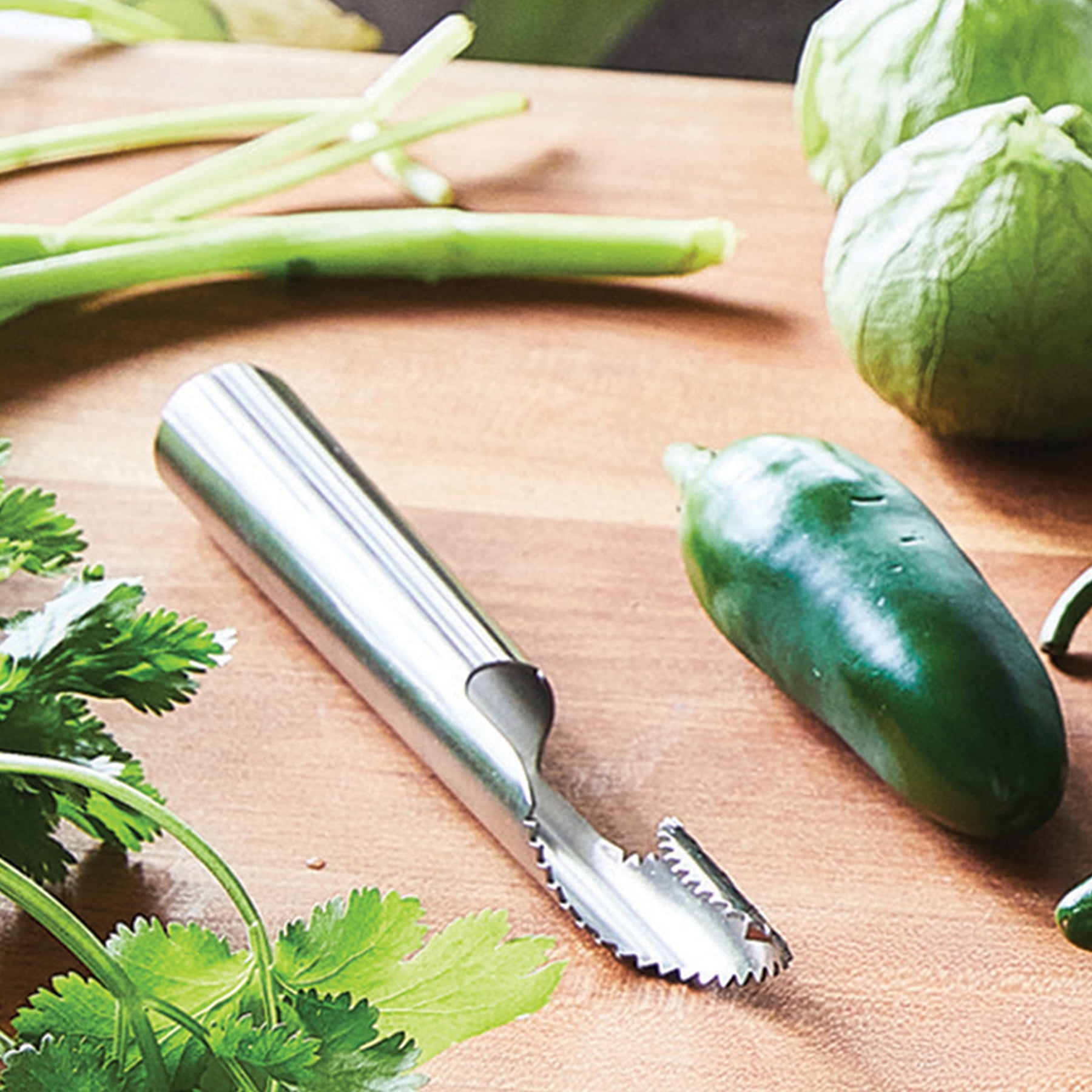 Stainless steel pepper de-seeder tool on a wooden surface surrounded by fresh vegetables and herbs.
