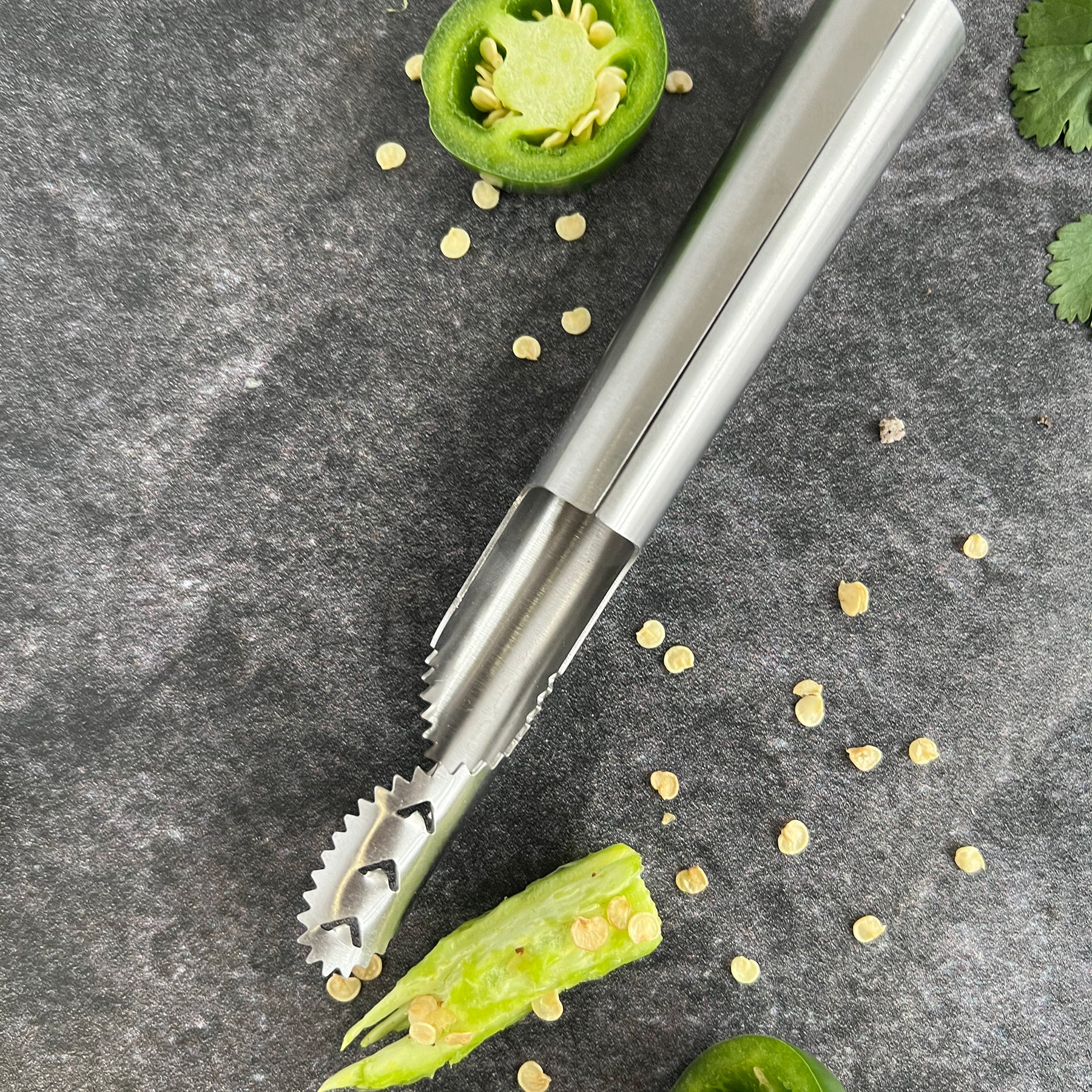 Stainless steel pepper de-seeder tool next to halved green pepper and scattered seeds on a dark surface.