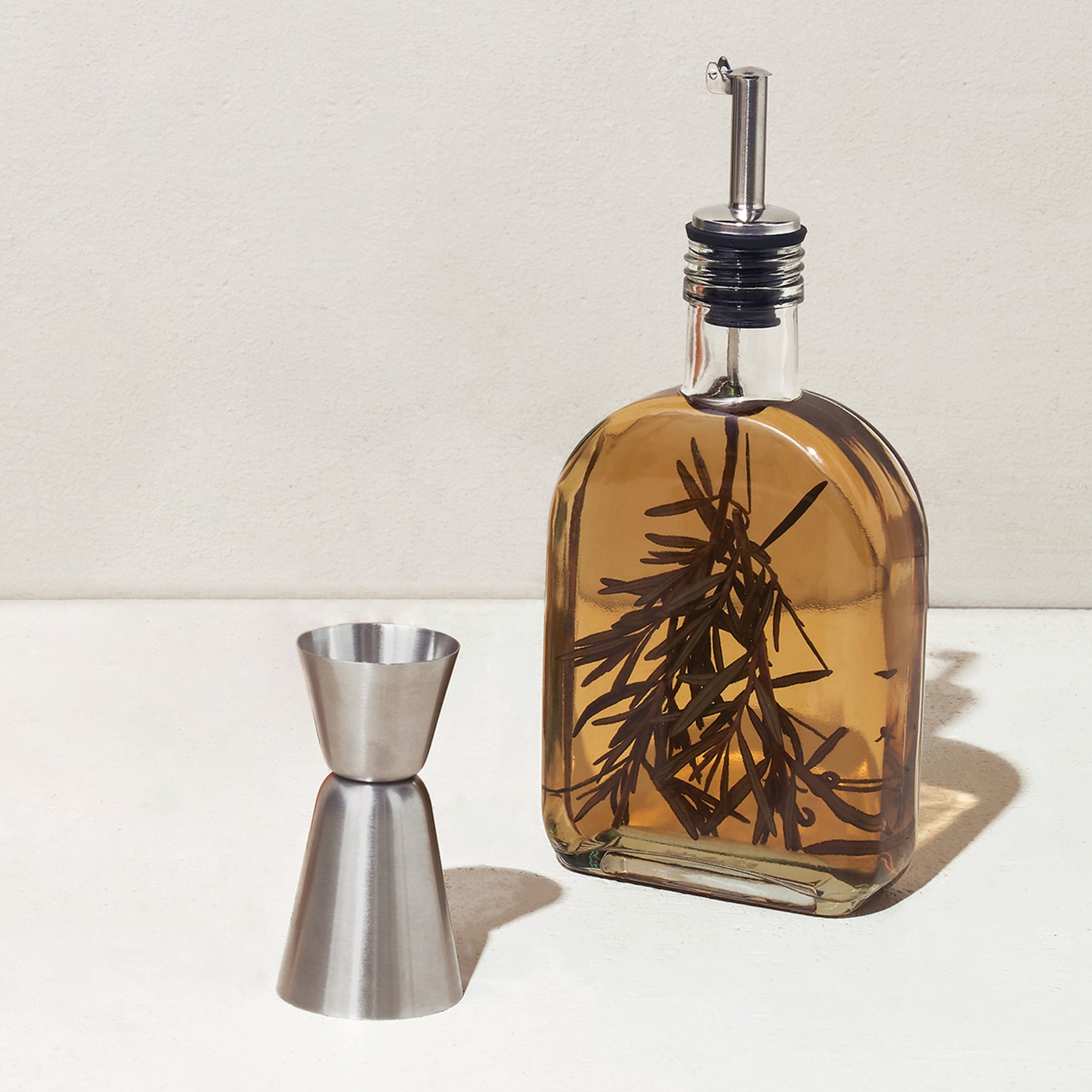 Infusion glass flask filled with infused liquid and herbs, accompanied by a stainless steel jigger on a light surface.