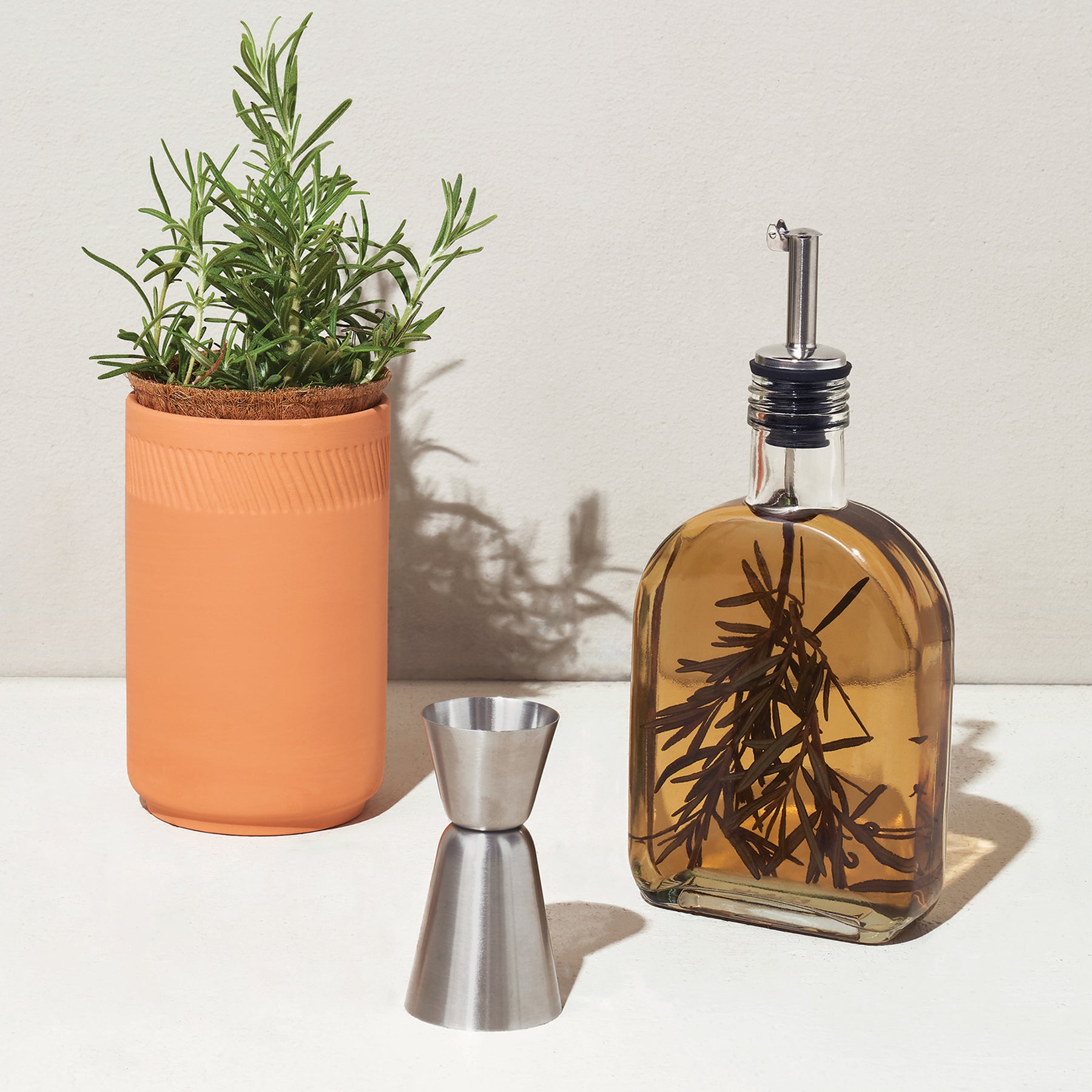 Infusion glass flask with rosemary, stainless steel jigger, and a terracotta planter with a green plant.