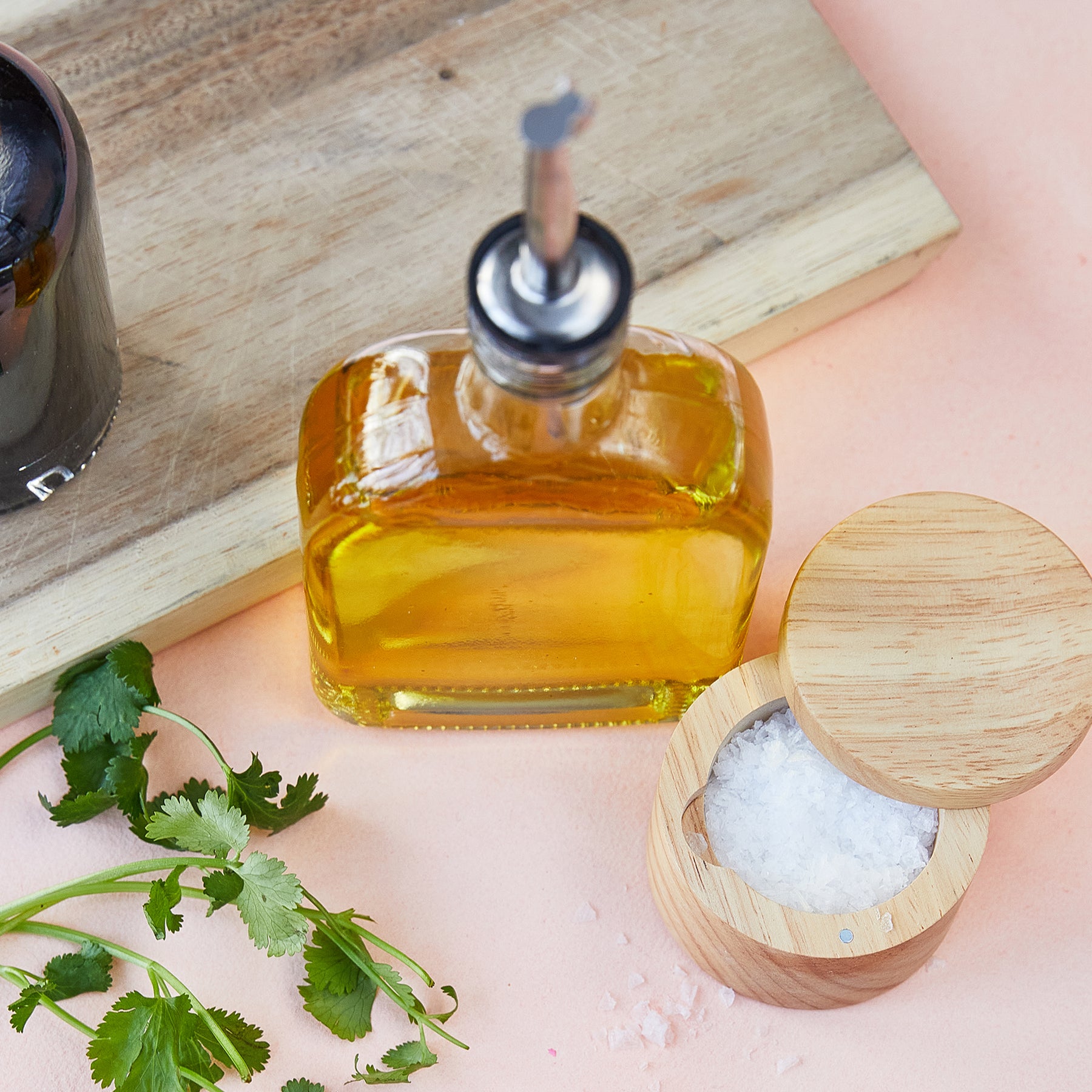 Infusion glass flask with oil, wooden salt container, and fresh cilantro on a pink surface.