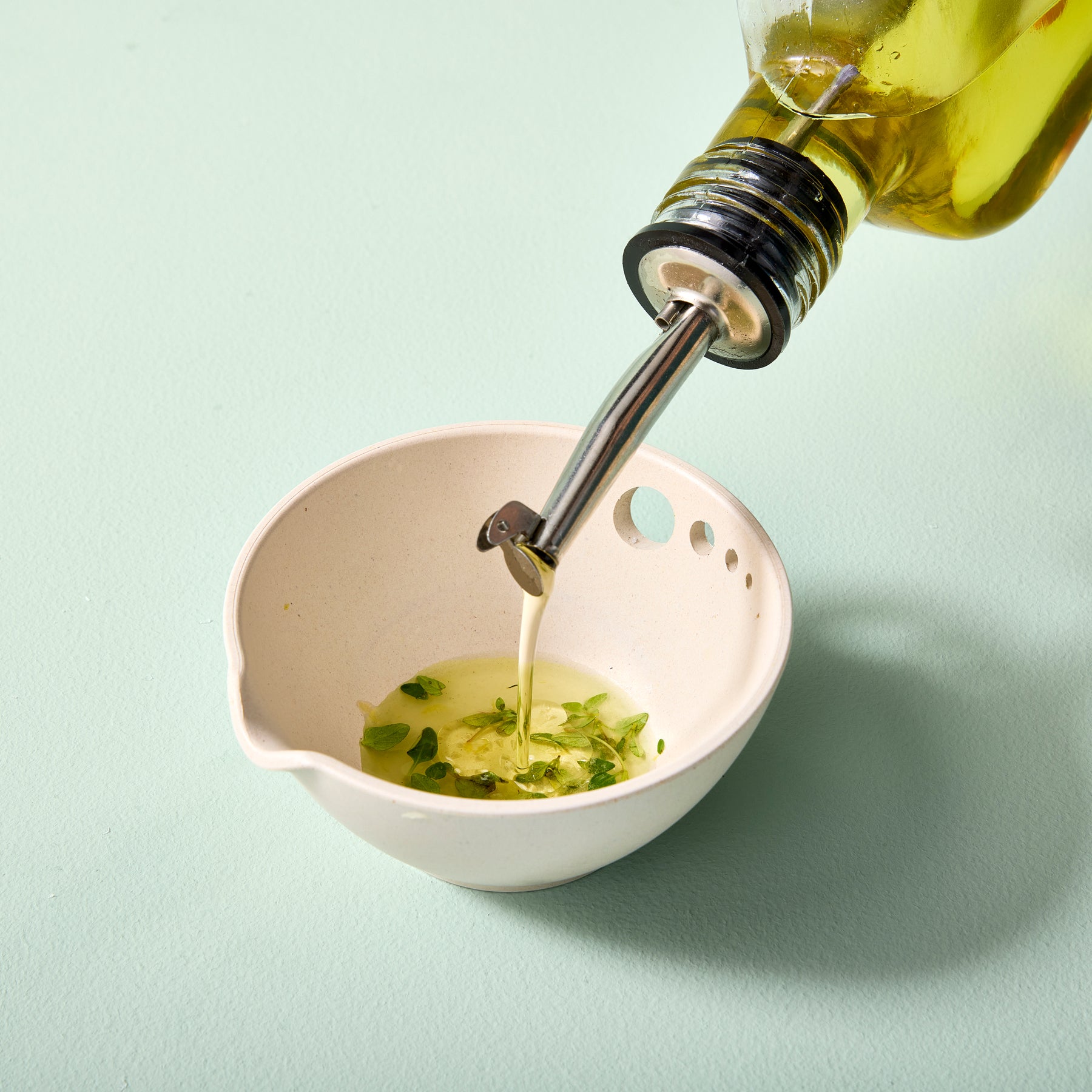 A glass flask pouring infused oil into a small white bowl with herbs, set against a light green background.