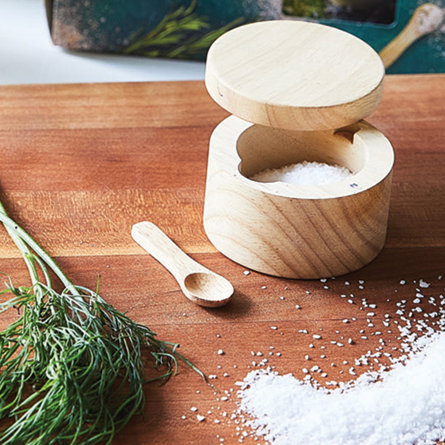 Wooden salt cellar with a lid and a small spoon, placed on a wooden surface with fresh herbs and scattered salt.