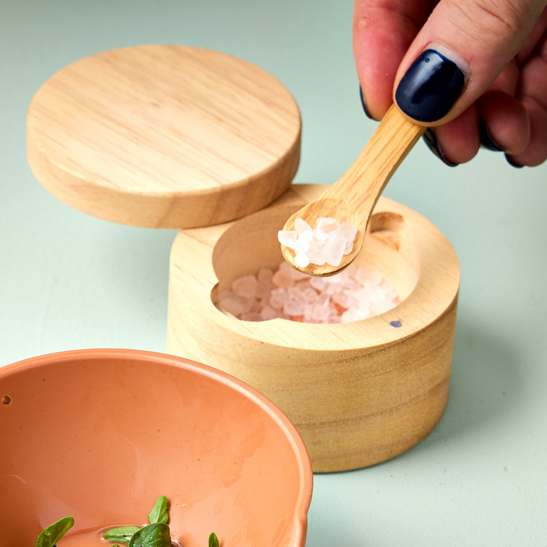 Bamboo salt cellar with a spoon, filled with pink salt, and a hand reaching in to scoop some out.