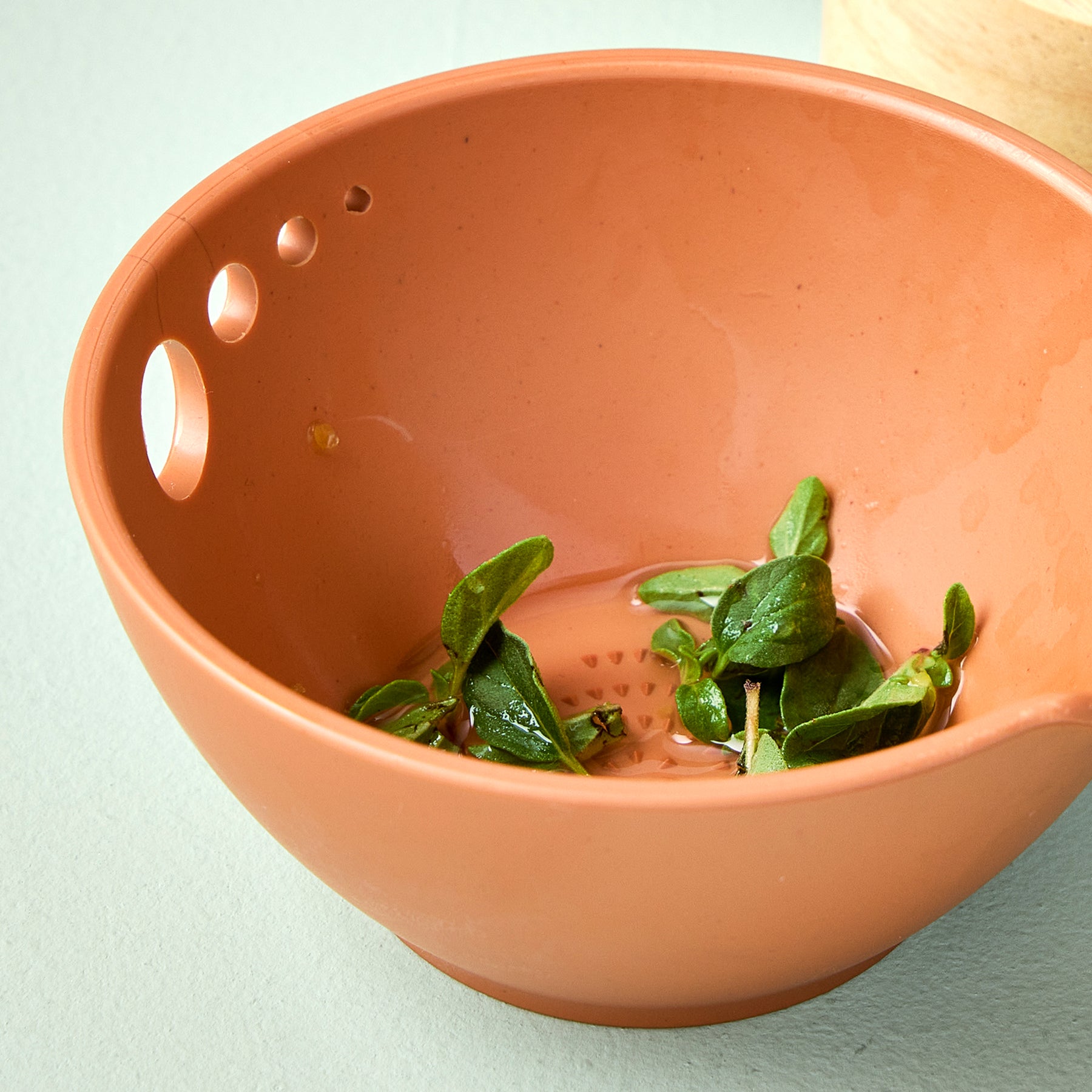 A terracotta-colored prep dish with drainage holes, containing fresh herbs and water, set against a light background.