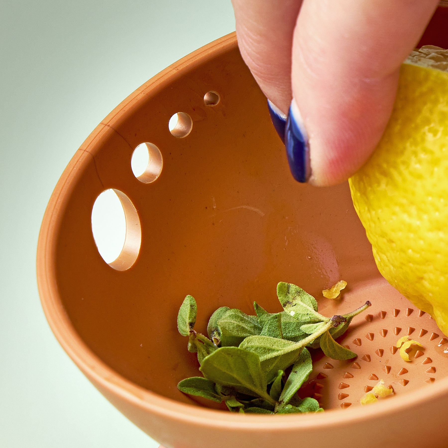 A hand holds a lemon over a brown dish with herbs and small holes for grating and peeling ingredients.